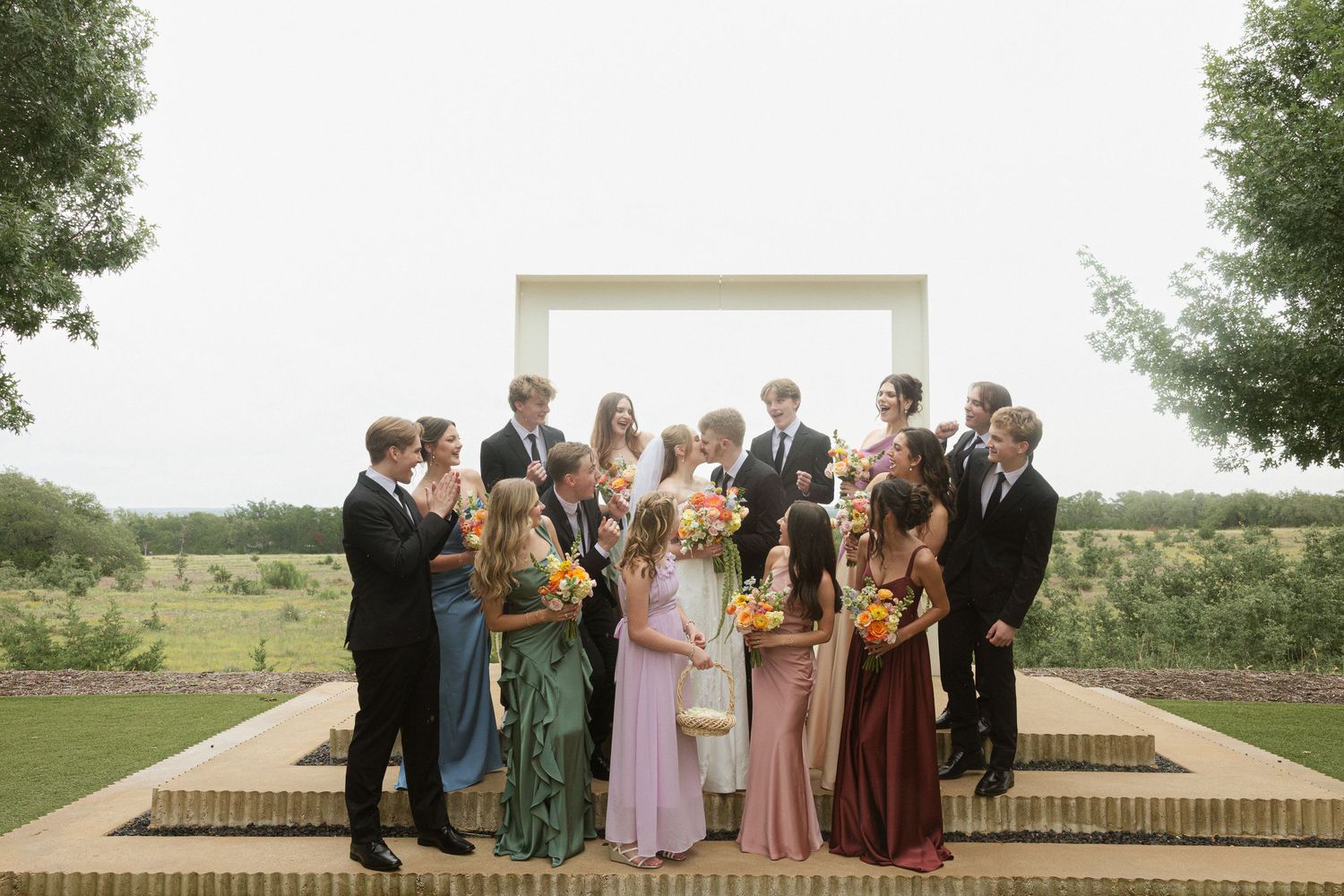 Wedding party group photo outdoors, featuring bridesmaids in colorful gowns and groomsmen in suits, gathered near a modern arch.