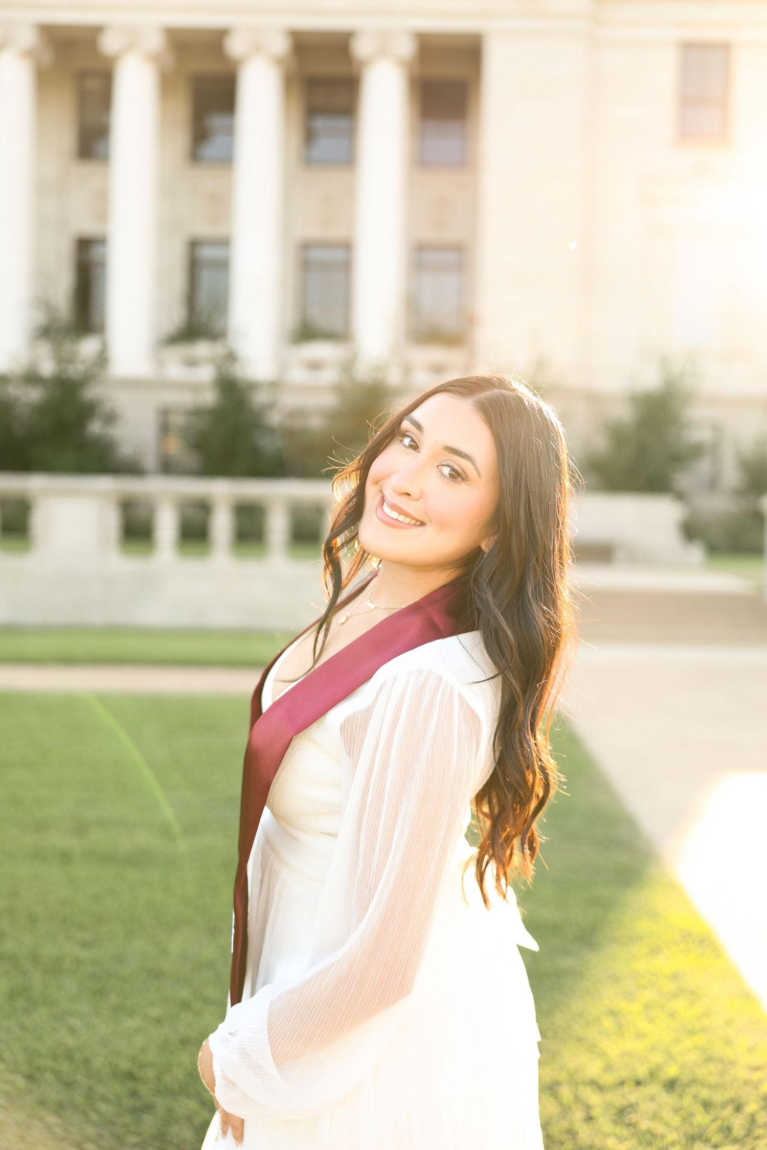 Graduate in white dress and maroon stole twirls joyfully in golden sunlight.