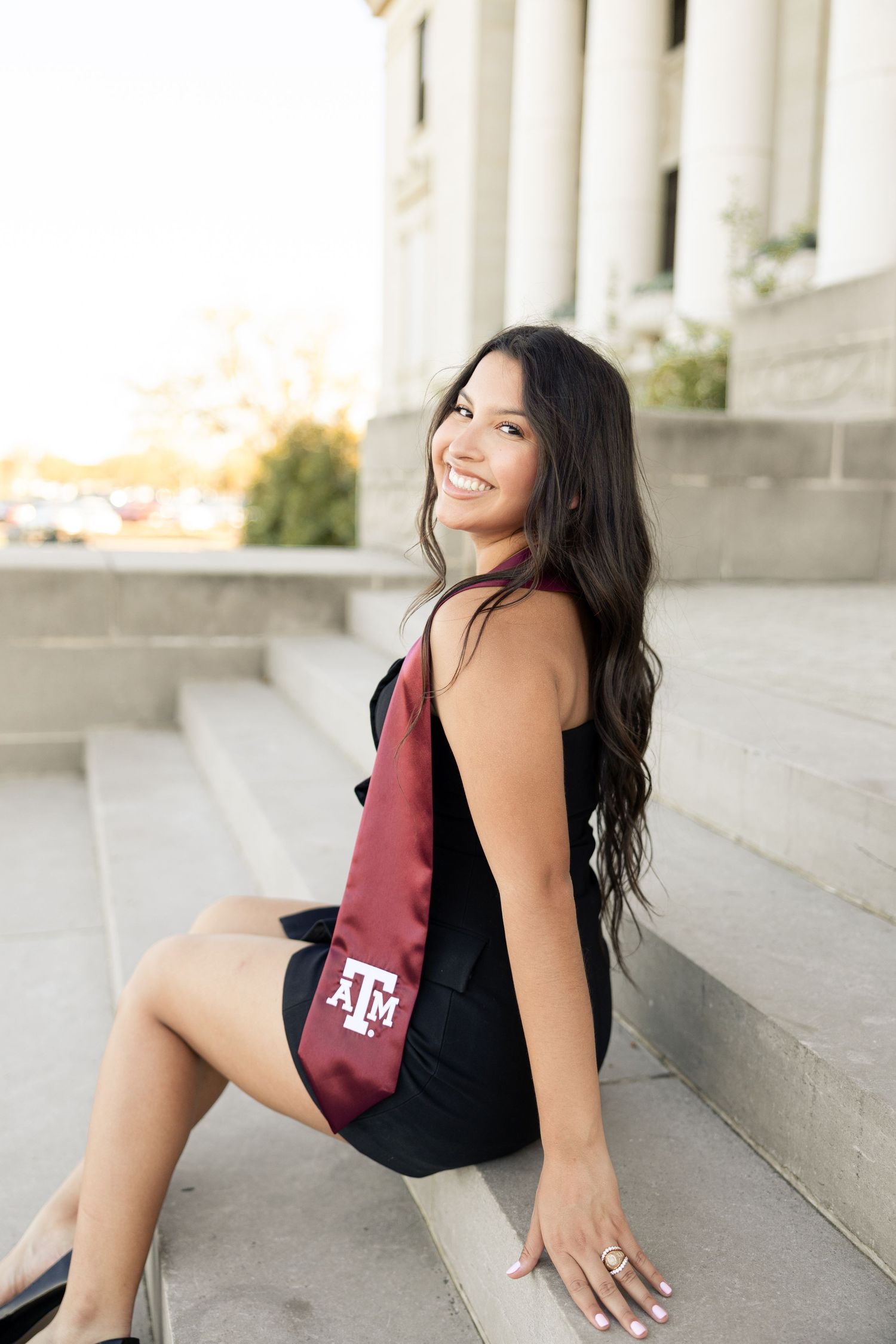 Graduate in black dress and maroon Texas A&M stole poses on stone steps during golden hour graduation photoshoot.