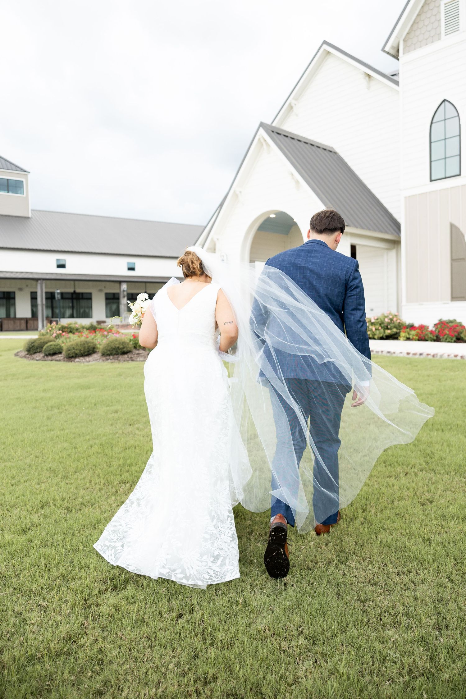 A couple walks away from a white church building with bridal veil floating in the breeze.