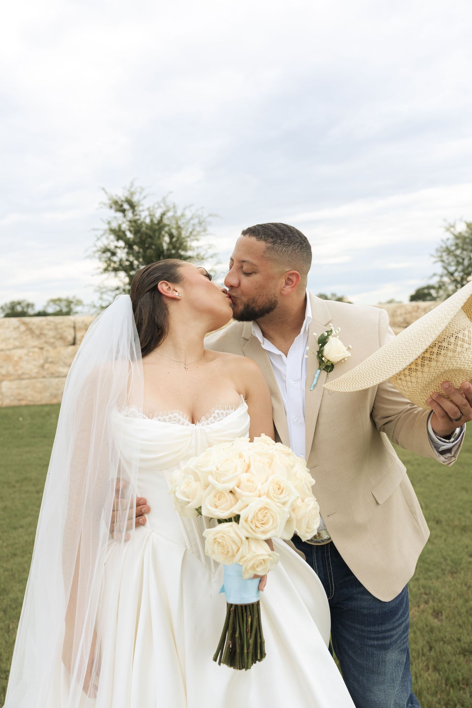 A couple sharing a romantic kiss on their wedding day with white roses and a scenic outdoor backdrop.