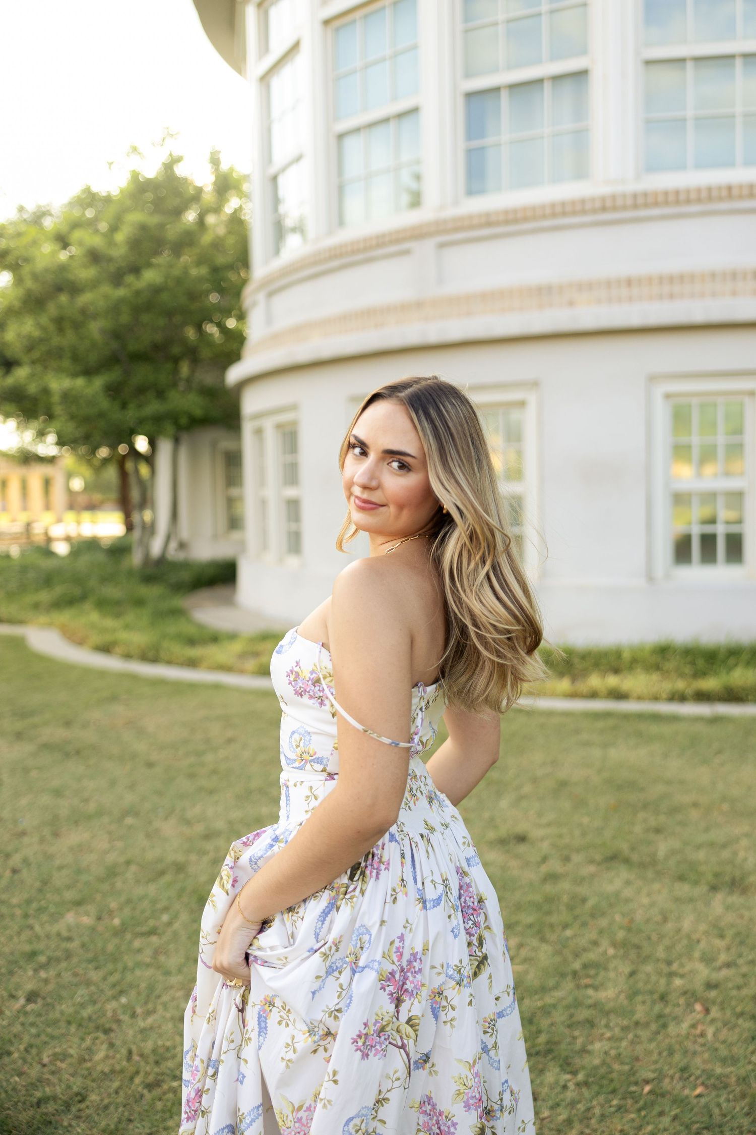 Woman in floral summer dress poses by curved white building during golden hour.