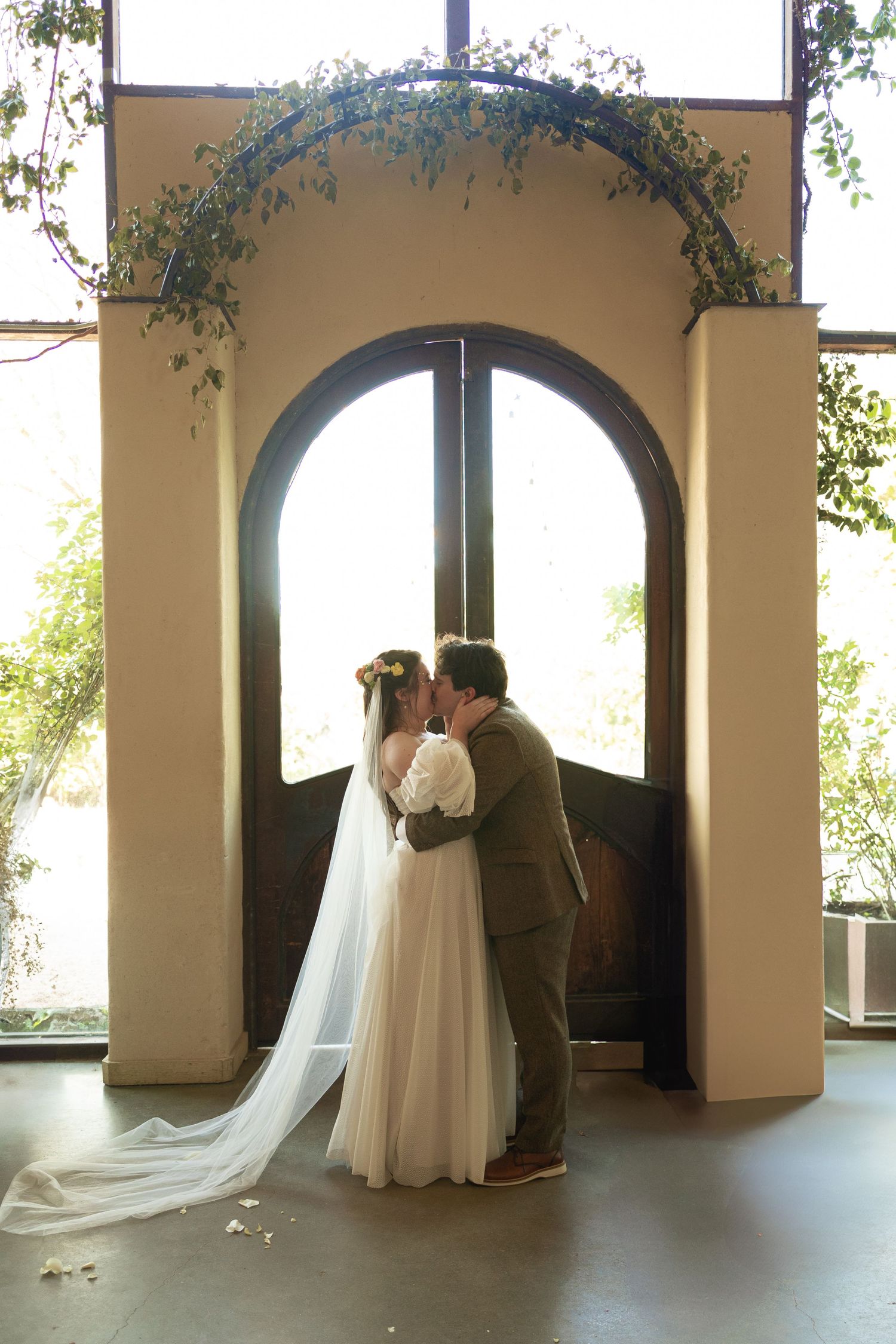 A romantic wedding moment under an archway decorated with greenery as light streams through glass doors.