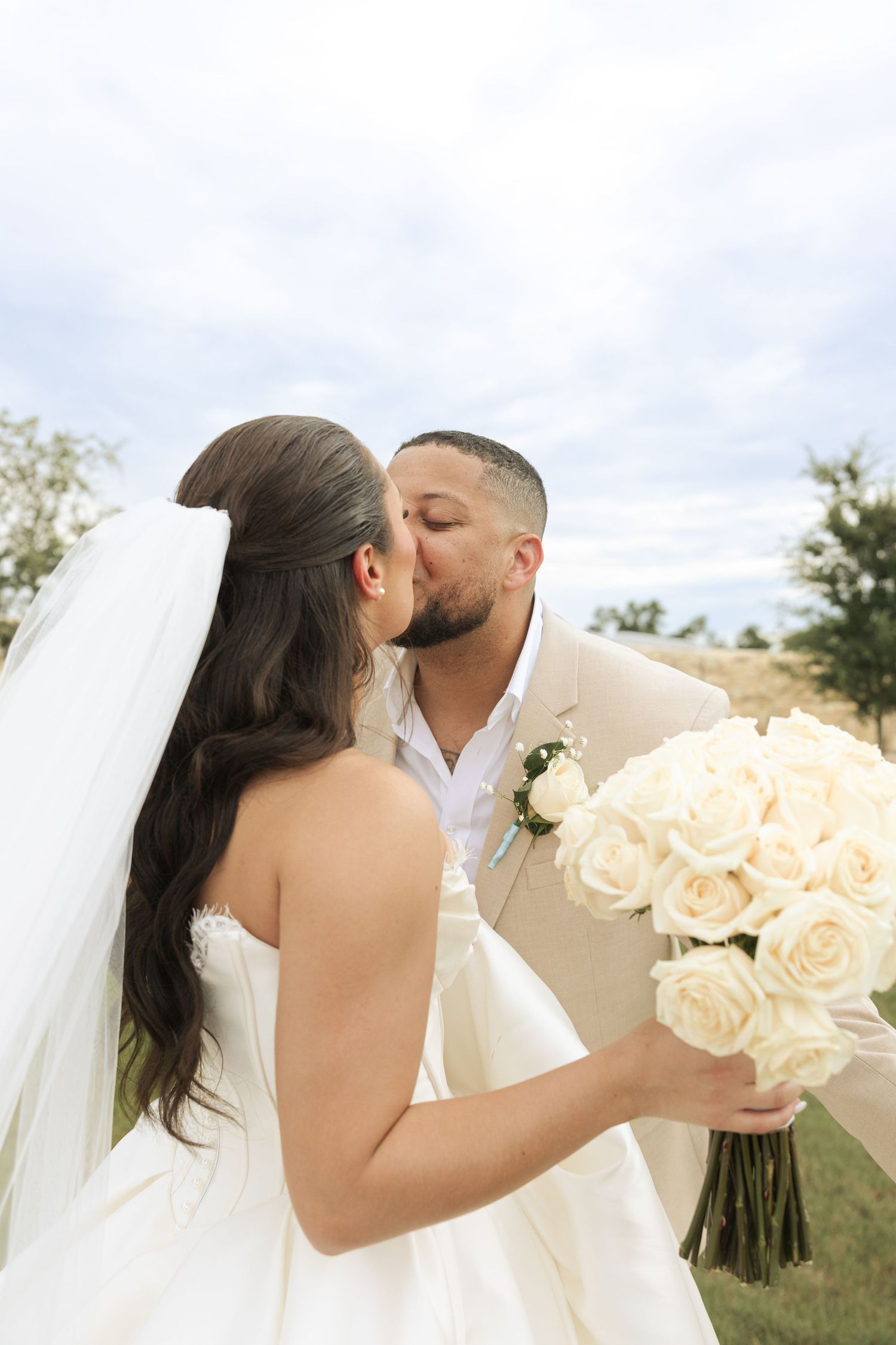 Romantic couple share a kiss while holding white rose bouquet during outdoor wedding ceremony.