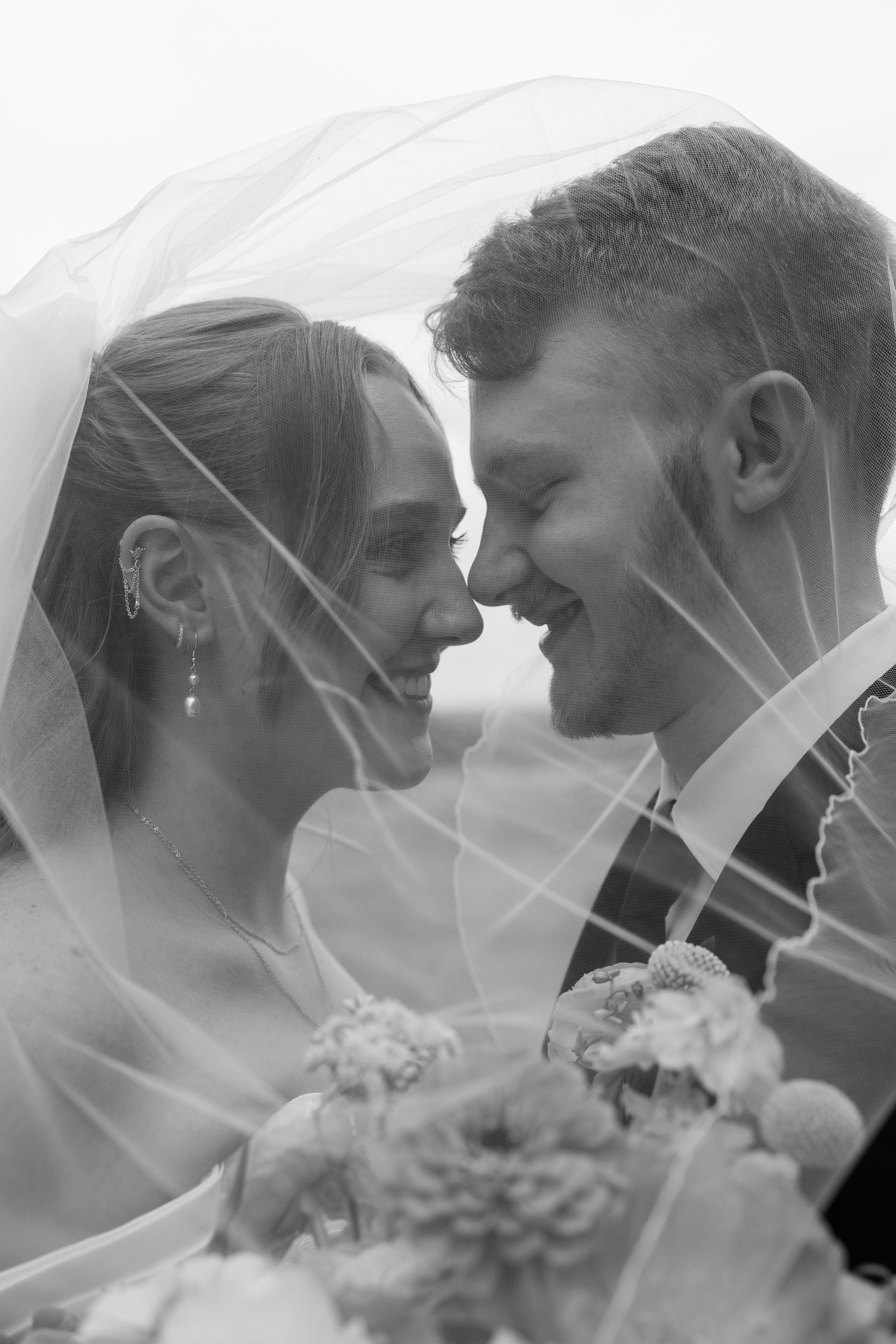 Black and white photo of smiling bride and groom under wedding veil, sharing an intimate moment with bridal bouquet.