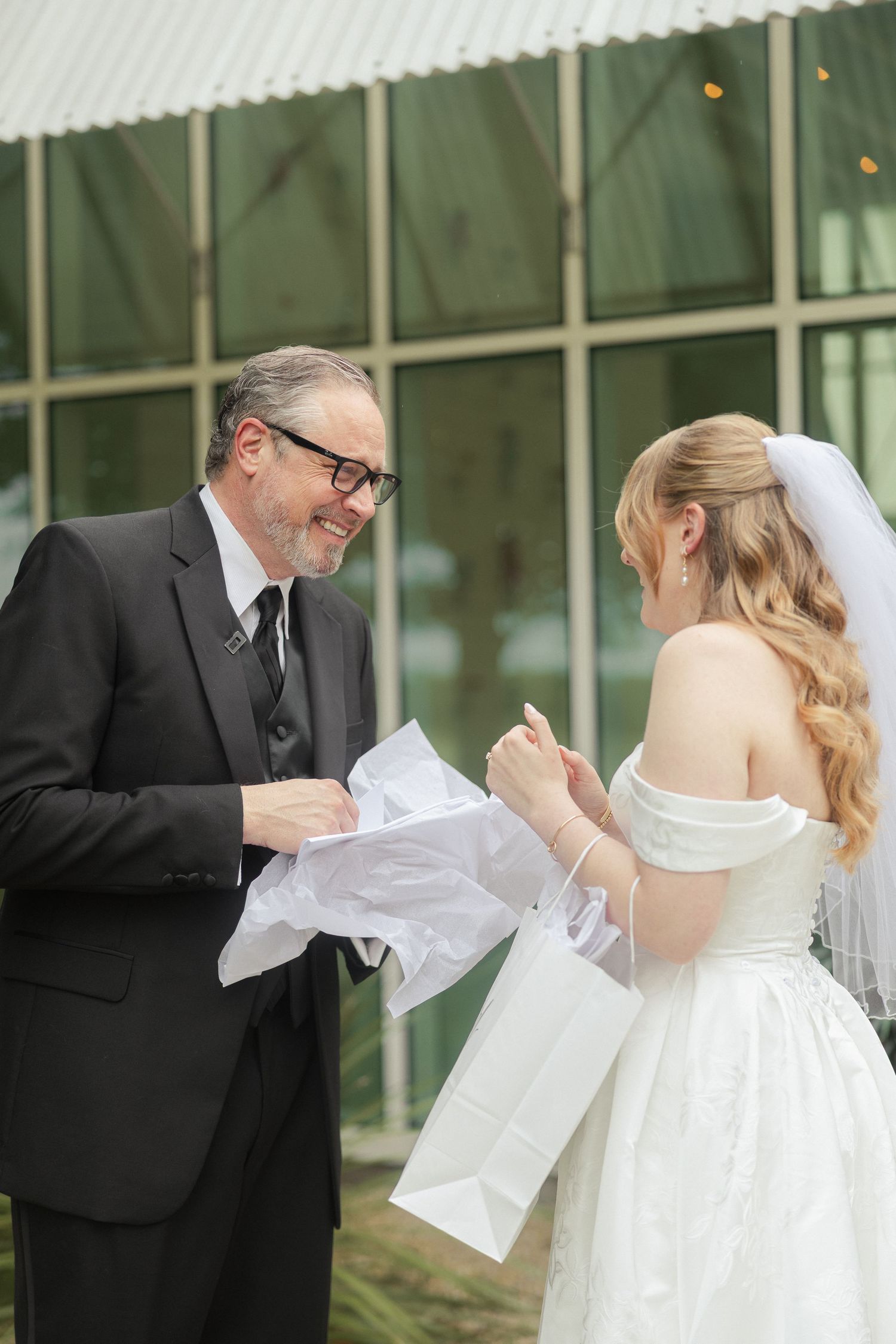 A bride in a white gown and veil receives a gift from a smiling man in a black suit outside a building.