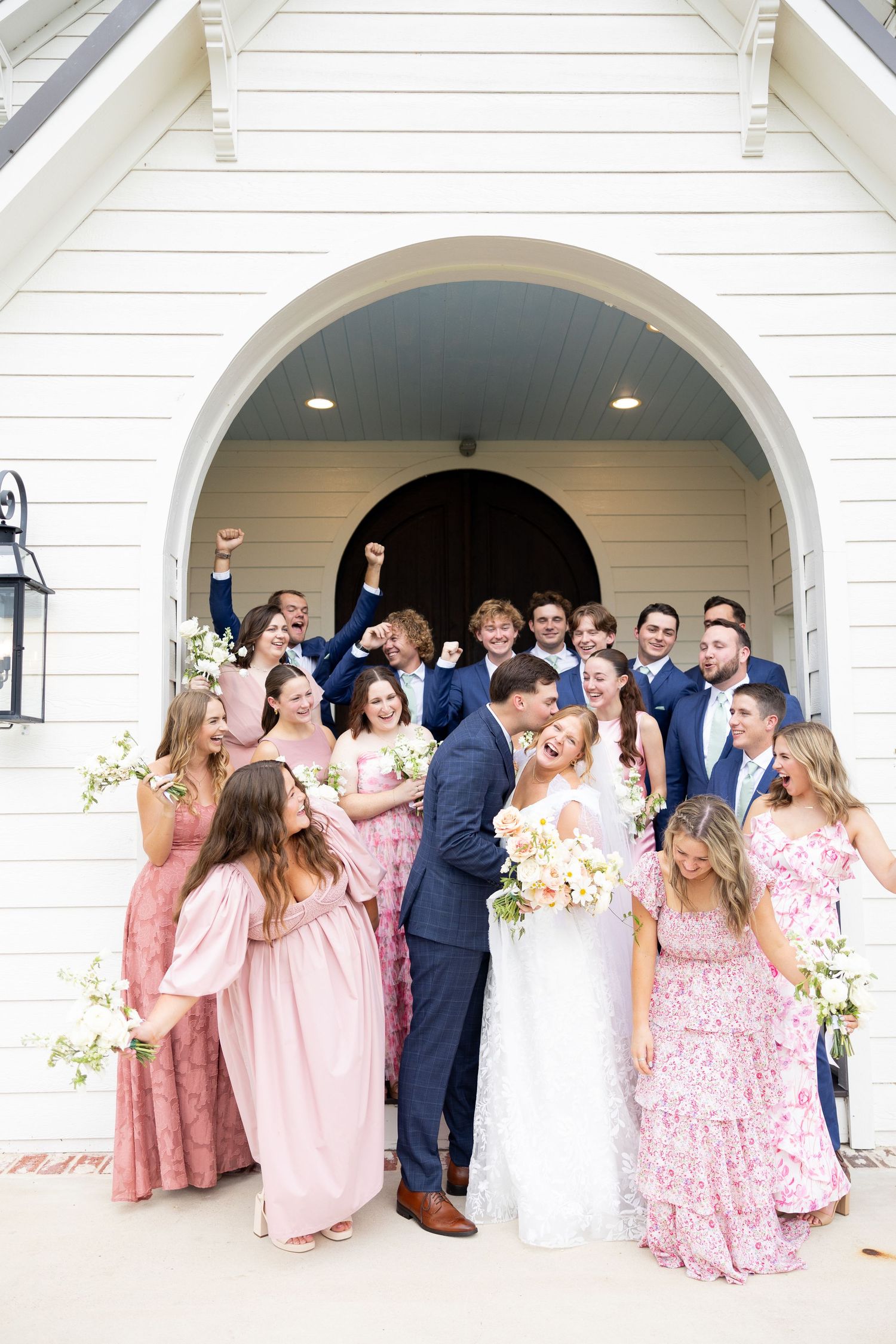 Wedding party poses under white archways wearing pink bridesmaid dresses and navy suits.