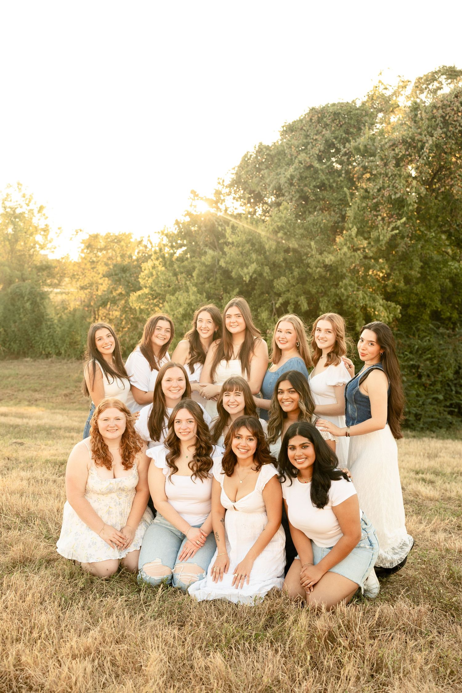 Group of friends in white tops sitting together on golden grass during sunset for casual outdoor photoshoot.