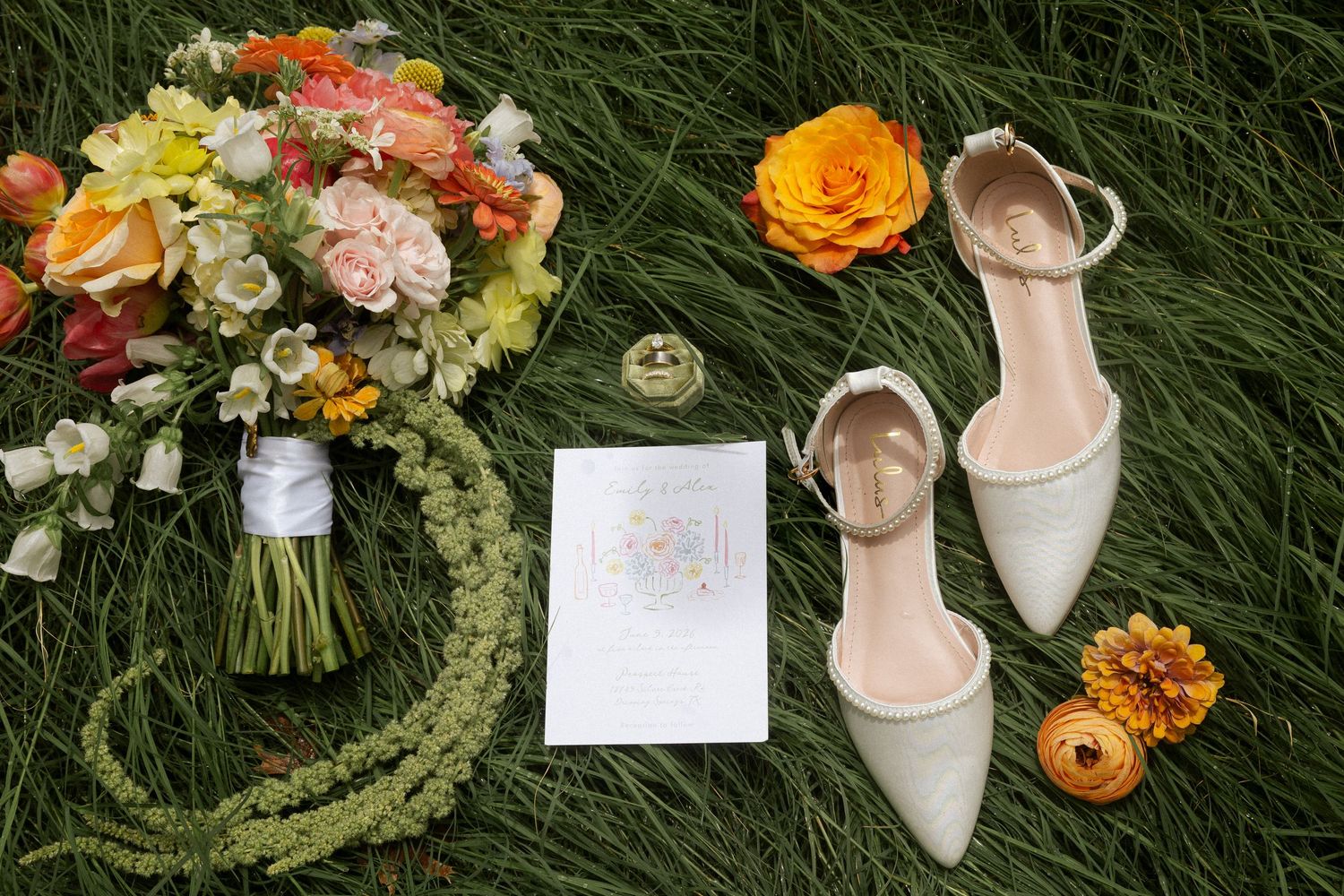 Bridal flat lay on grass featuring colorful flower bouquet, sage green heels, wedding invitation, ring, and orange floral accents.