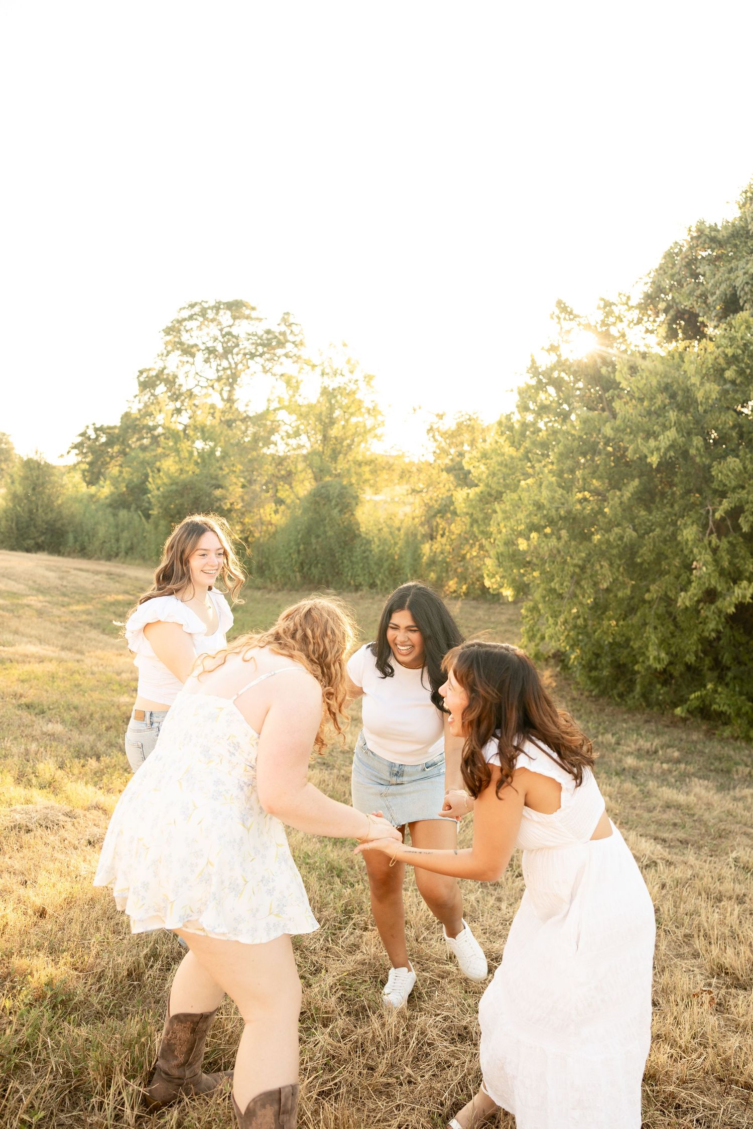 Women in white dresses dancing joyfully together in a sun-lit field.