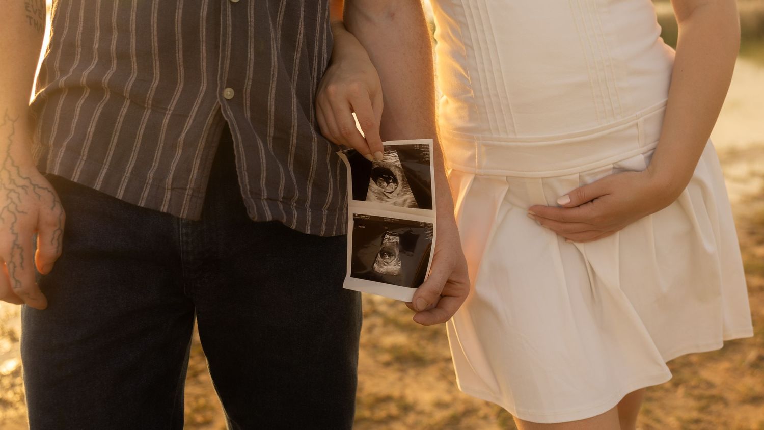 A couple holds an ultrasound photo while standing together outdoors at sunset.