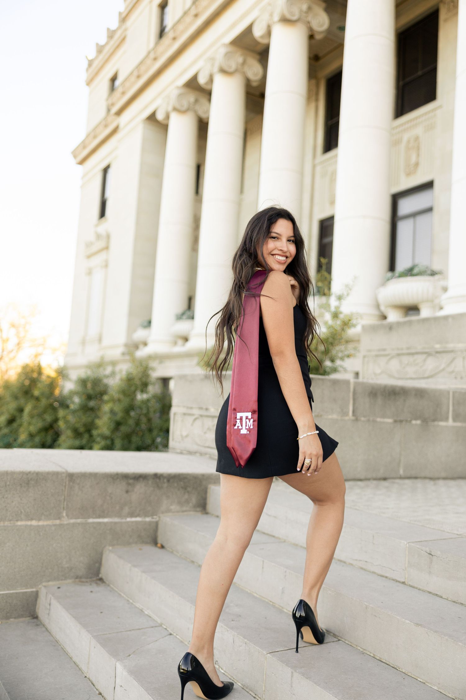 Graduate wearing black dress and maroon stole walks up classical stone steps during photo session.