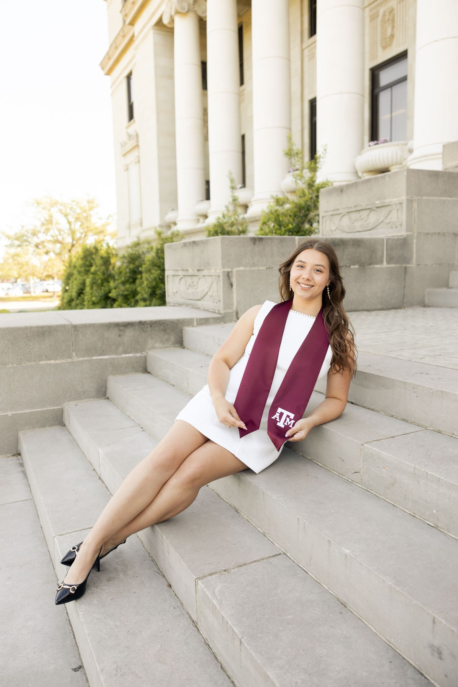 Graduate in white dress and maroon stole poses on stone steps of columned building.