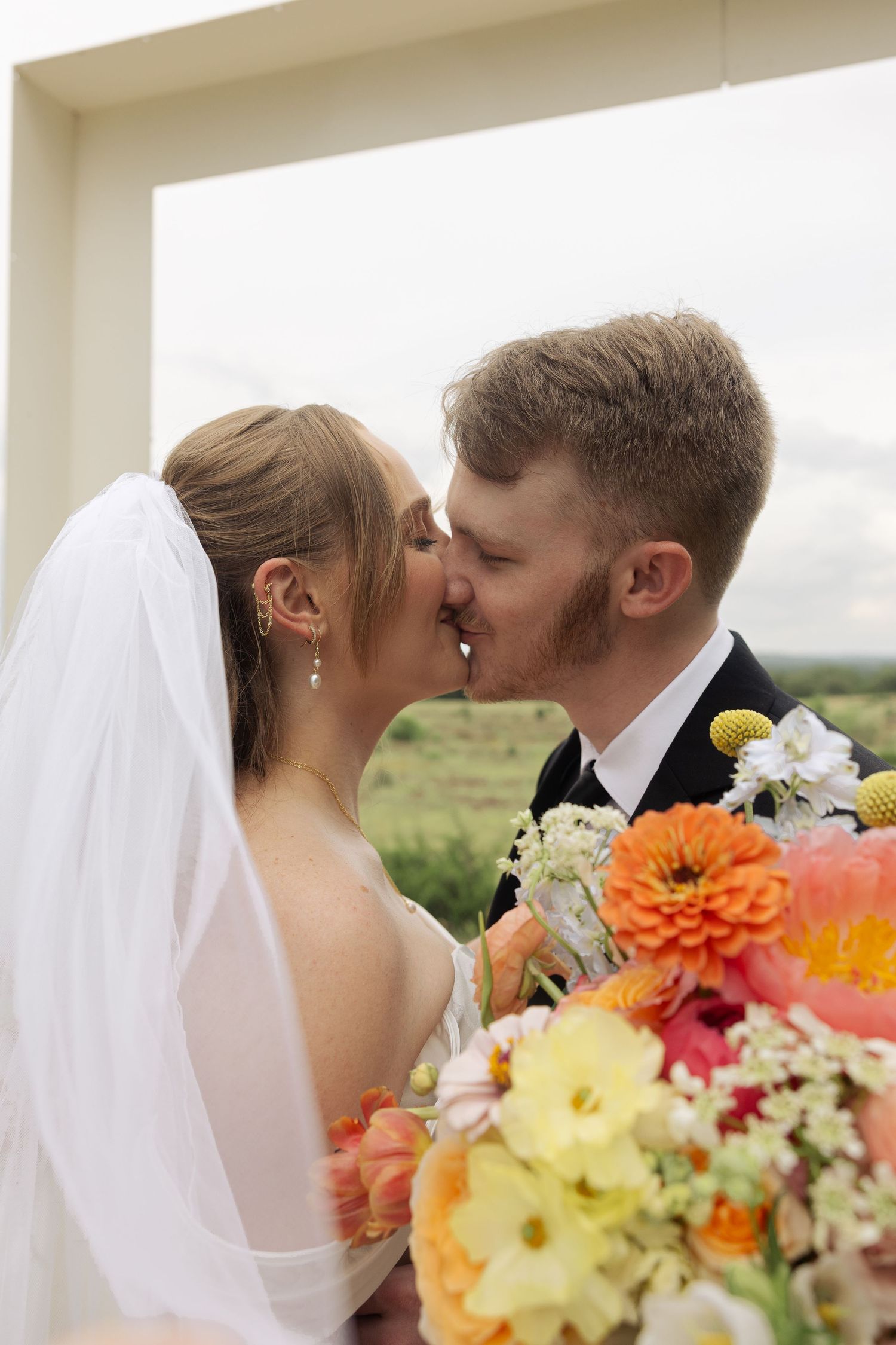 Bride and groom sharing a kiss at their wedding ceremony, with a vibrant colorful bouquet in the foreground.