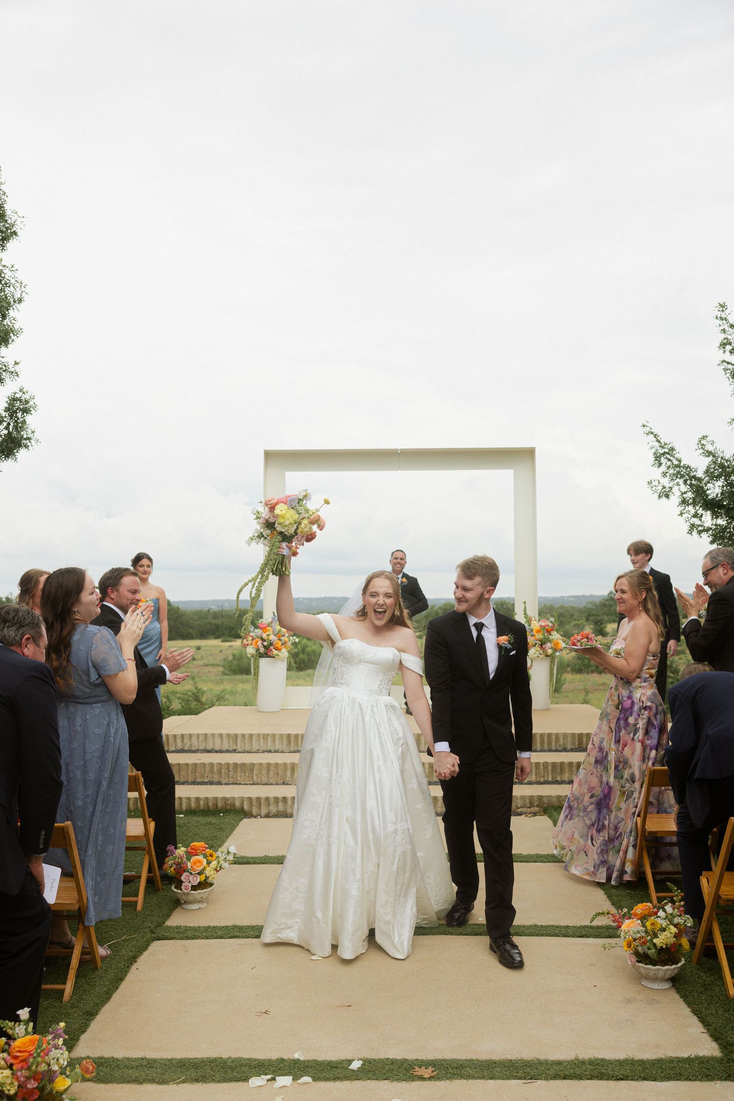 Joyful bride raises bouquet while walking down aisle with groom at outdoor wedding ceremony.