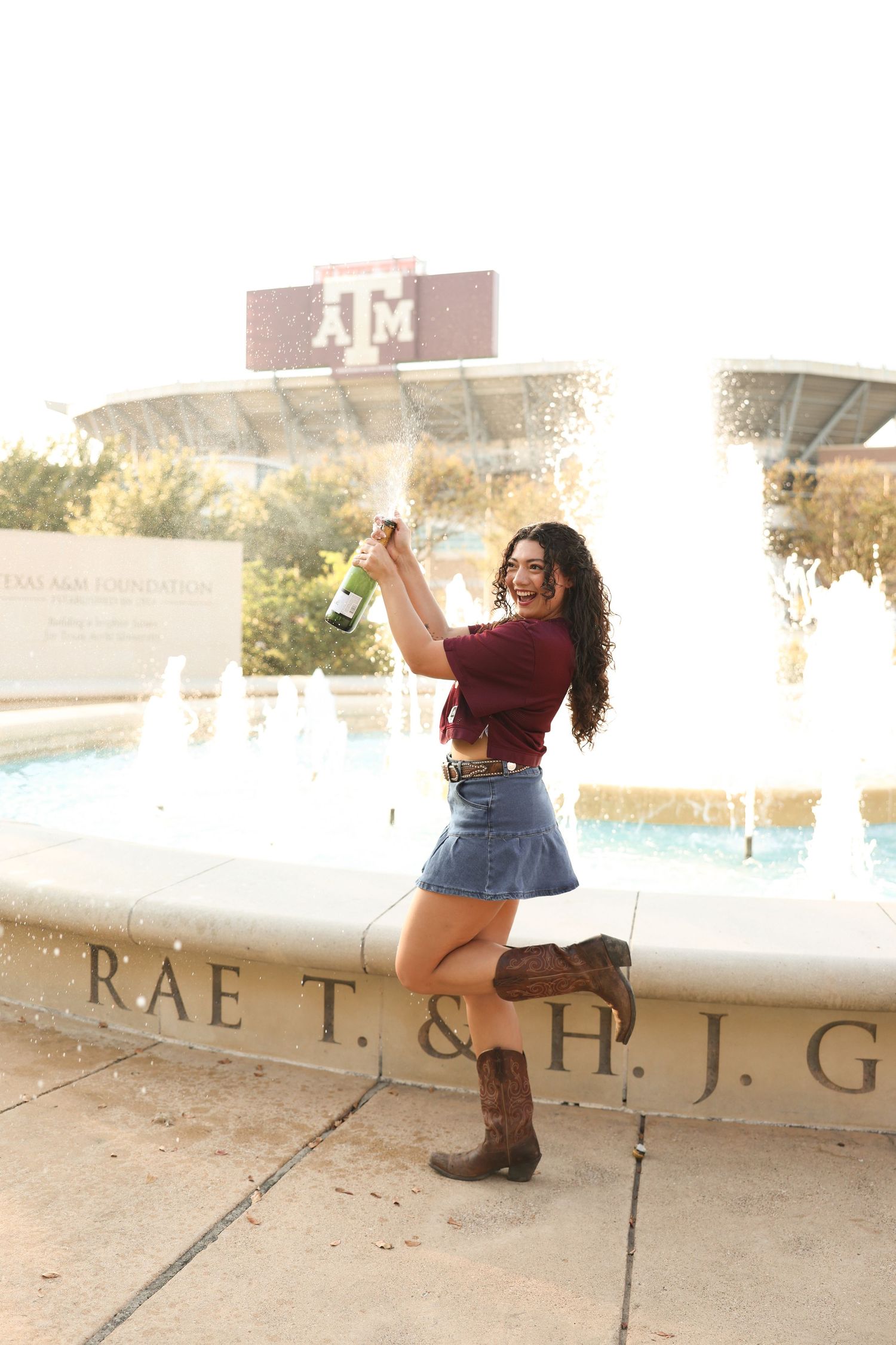 Graduate in casual denim and maroon top poses near campus signage.