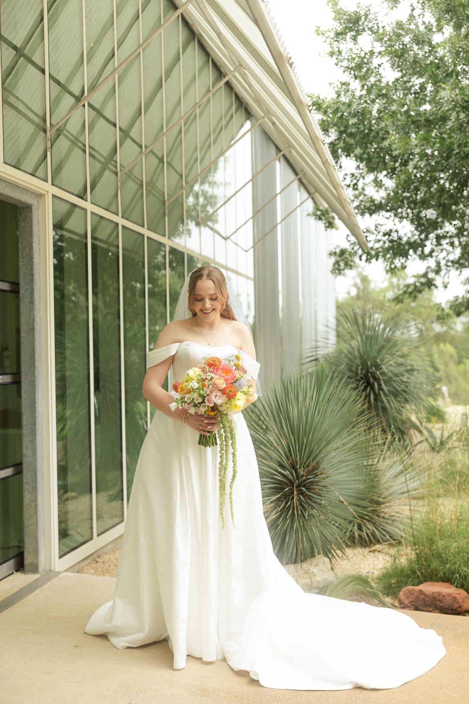 Bride in off-shoulder white gown holding colorful bouquet, smiling outside a greenhouse venue with desert plants.