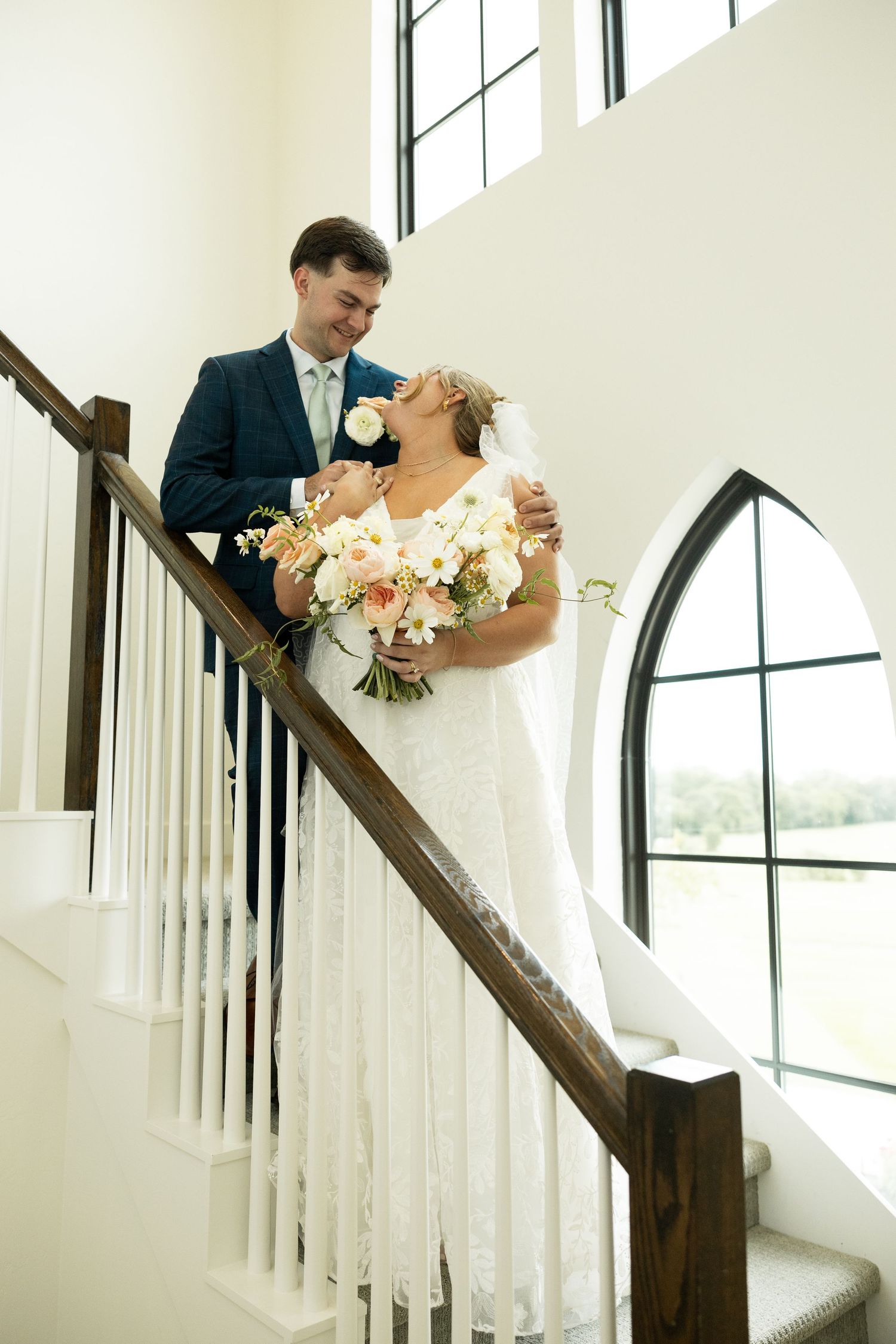 A couple poses on an elegant white staircase in a chapel with gothic windows.