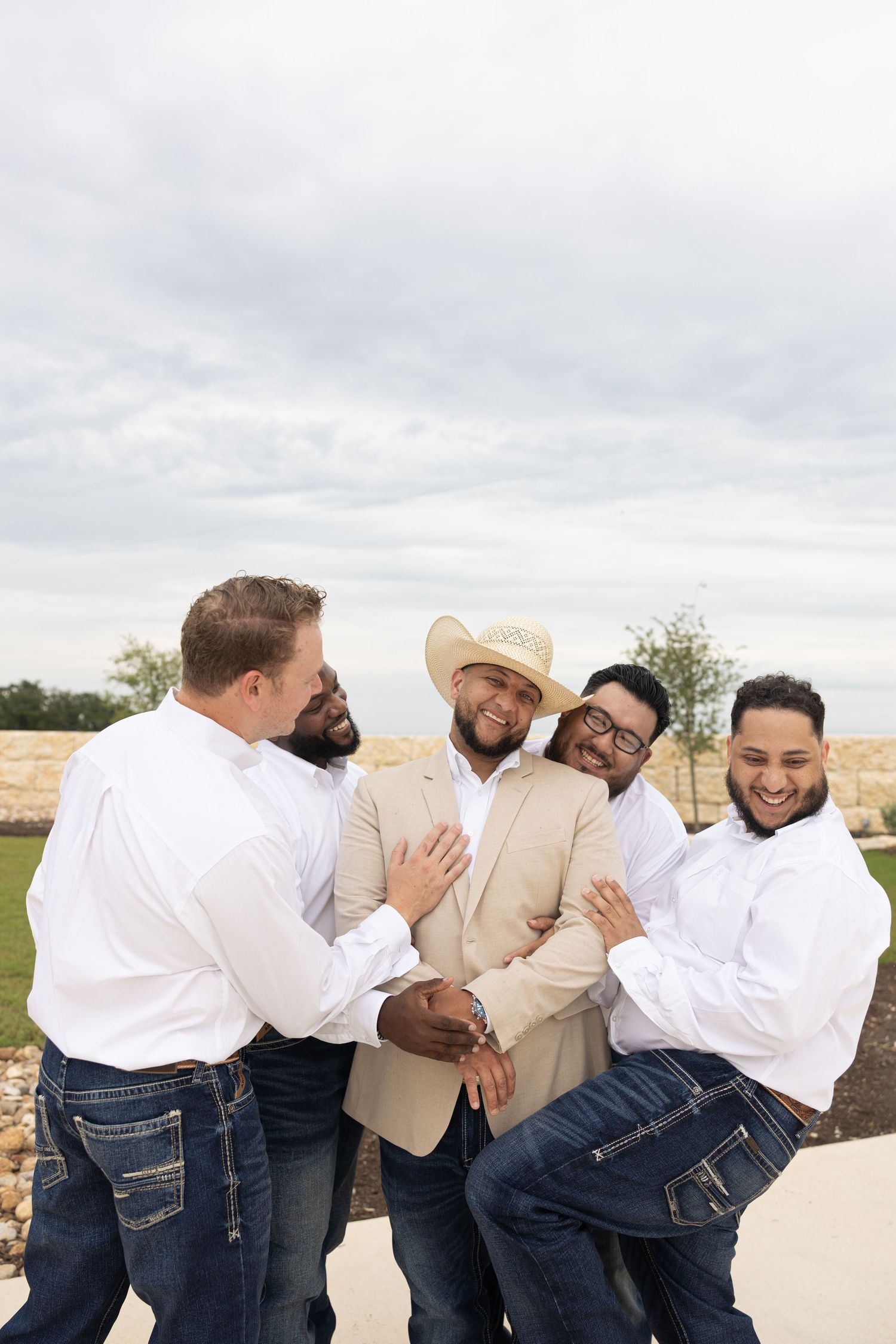 Groomsmen in white shirts and jeans share a playful moment with the groom in a beige blazer and cowboy hat.