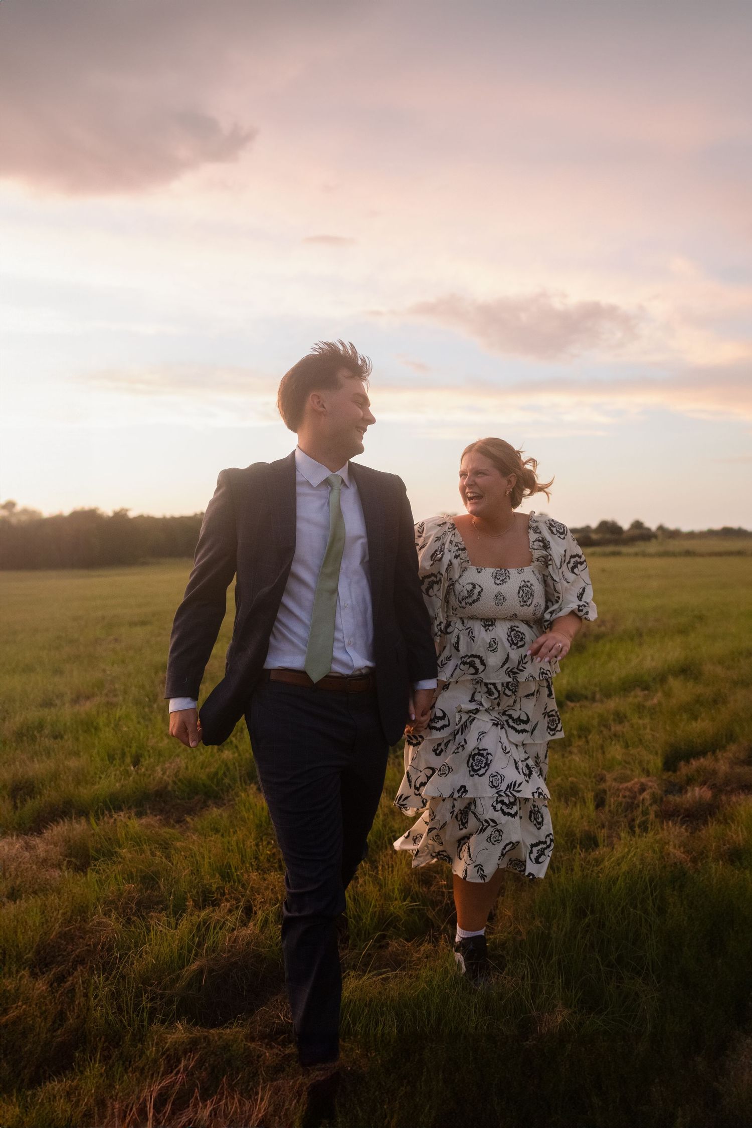 Couple walks together through sunset-lit field in formal wear.