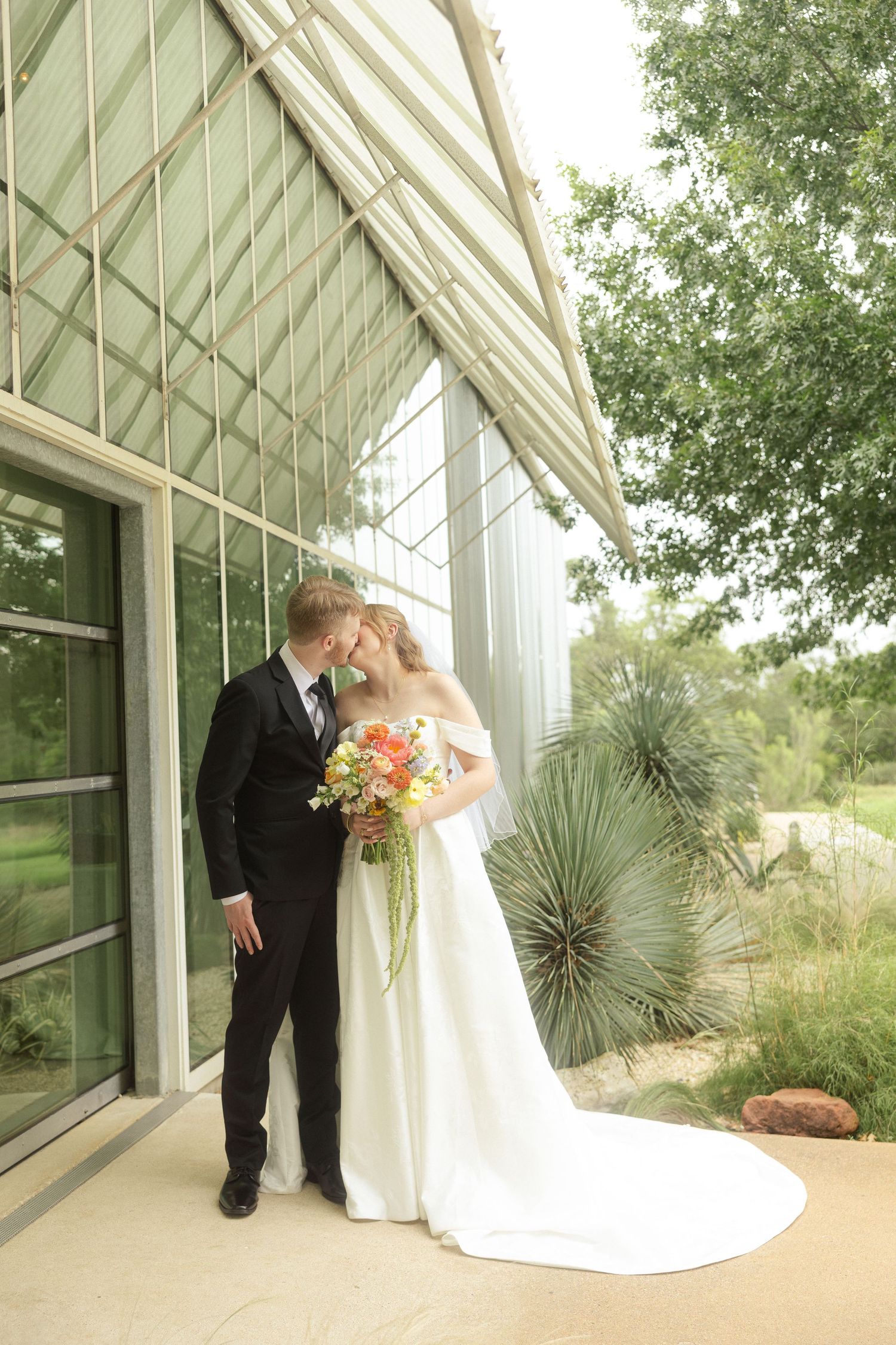 Bride and groom share a kiss outside a modern glass building, bride holding a colorful bouquet with long train.