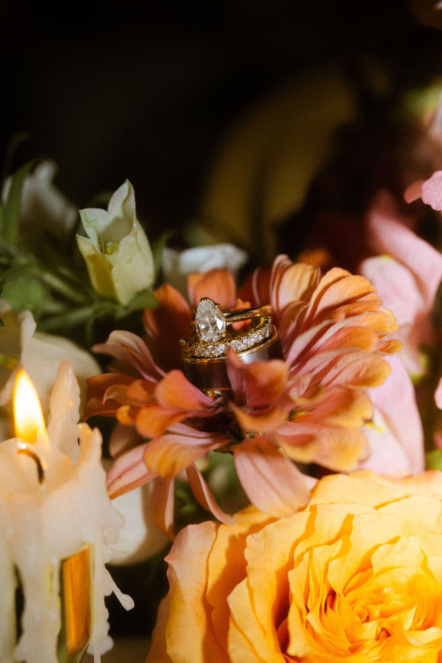 Gold diamond engagement ring and wedding band resting on pink flowers with candles and yellow roses in the background.