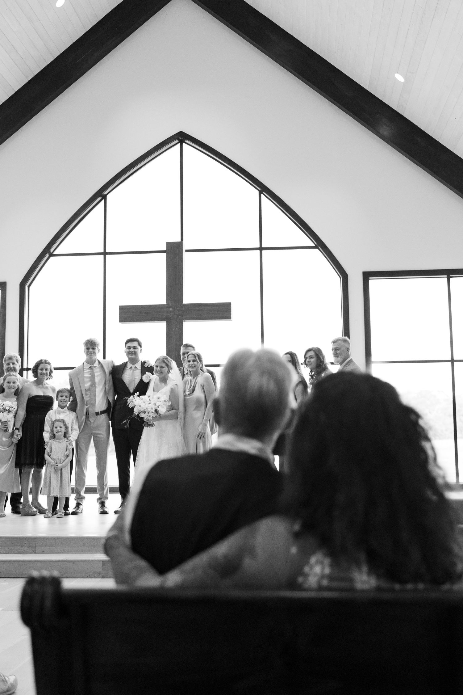 A church wedding ceremony with guests watching from wooden pews under a dramatic arched window with cross.