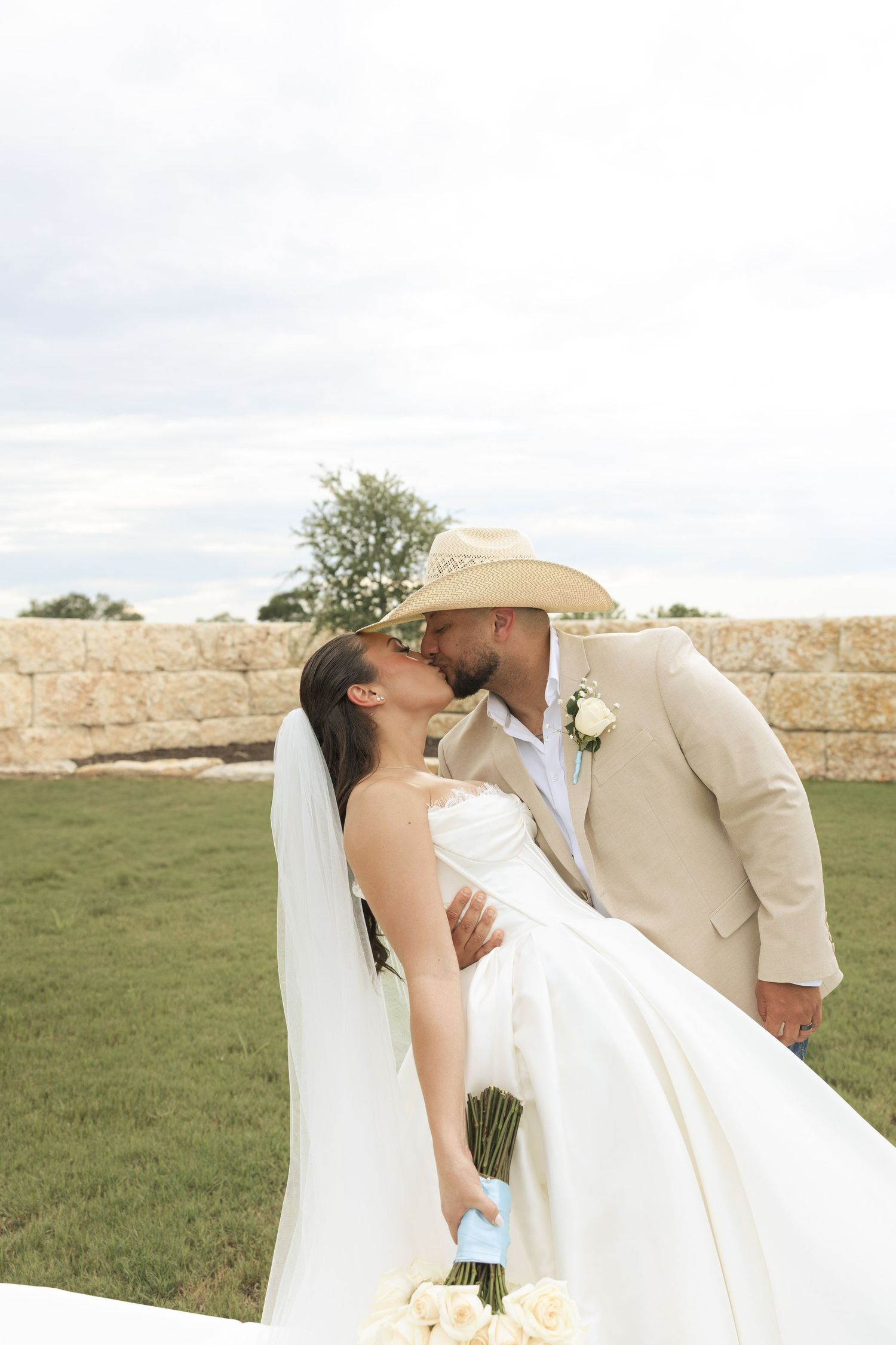 A romantic wedding couple shares a passionate kiss against a stone wall backdrop outdoors.