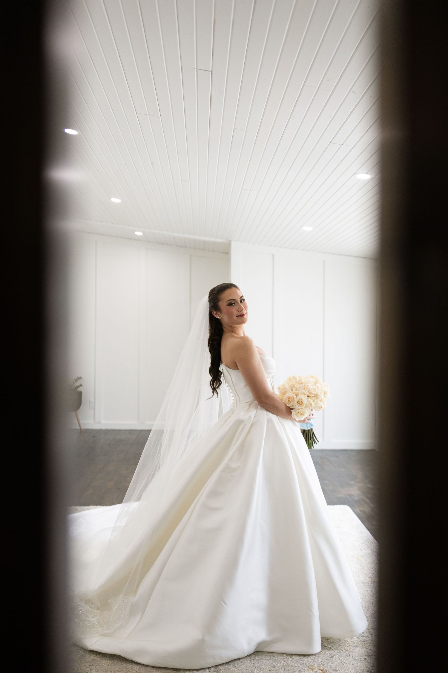 A bride in a luxurious white ballgown wedding dress poses elegantly with a cream bouquet in a bright room.