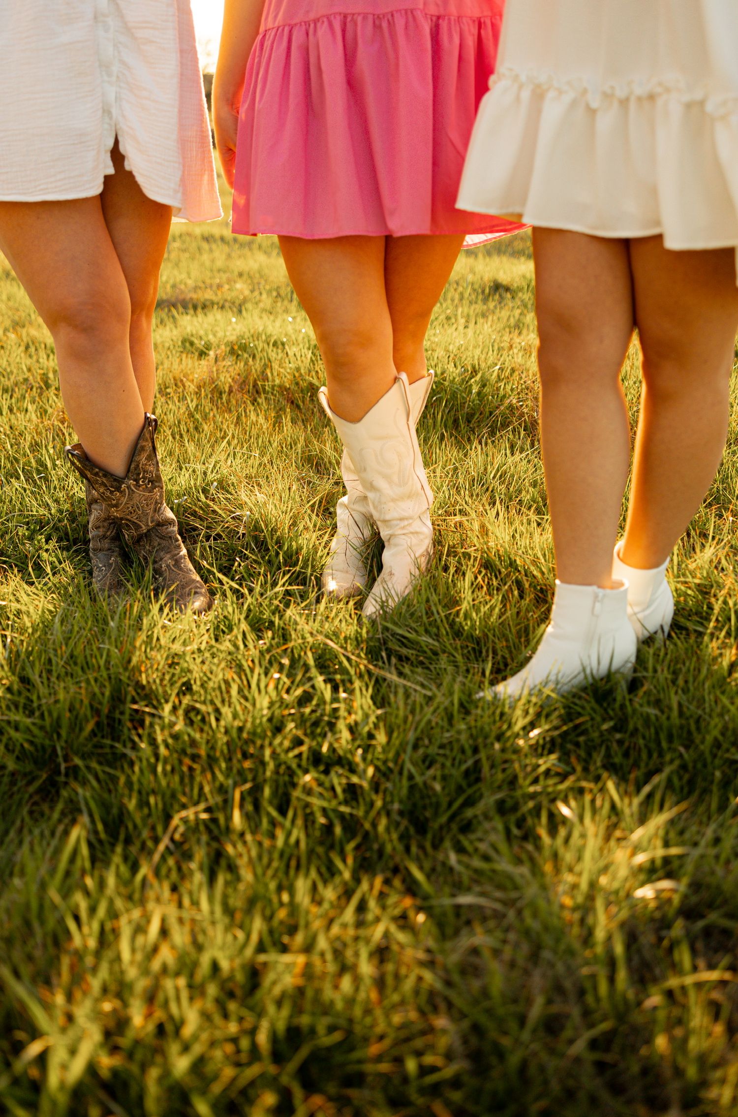 Row of legs showing colorful dresses and white boots against green grass at sunset.