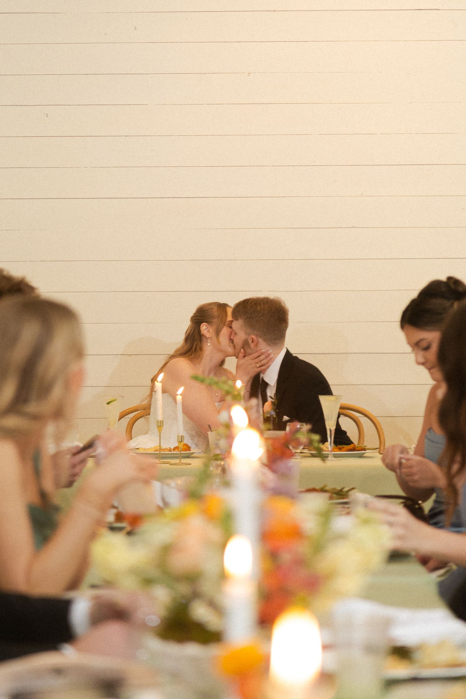 Bride and groom share a kiss at candlelit wedding reception dinner table surrounded by guests.