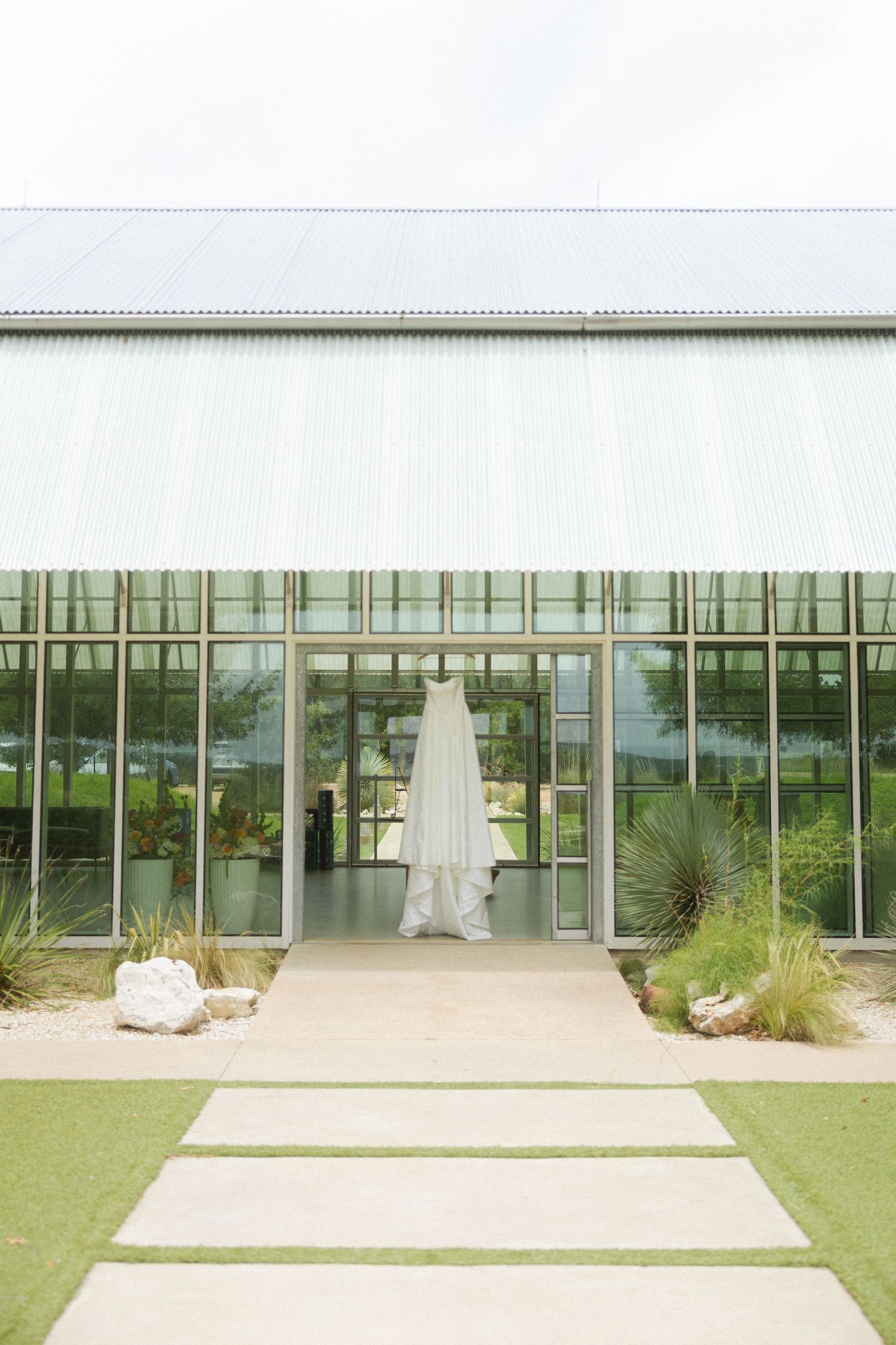 White wedding dress hanging at entrance of modern glass greenhouse venue with lush greenery.
