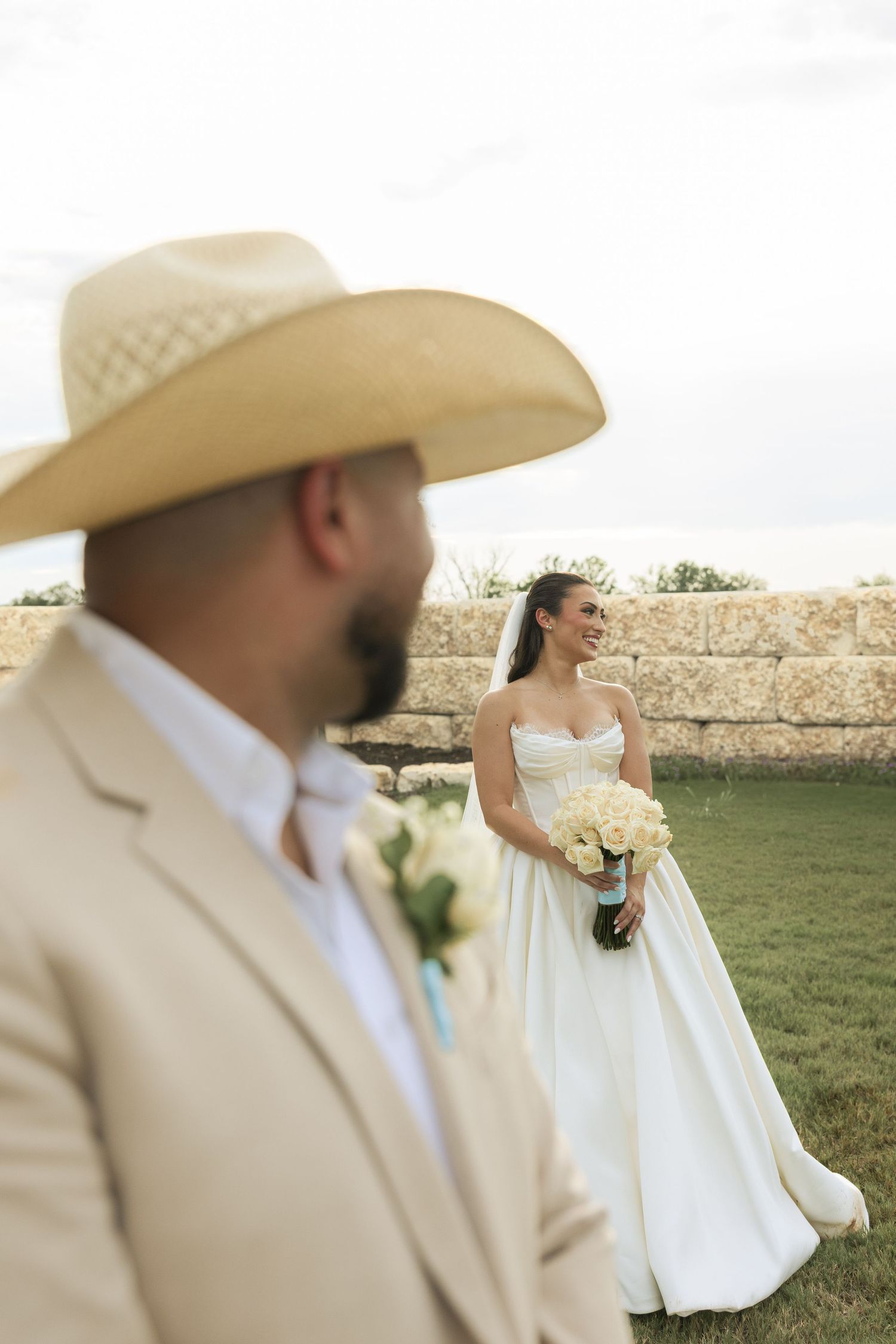 Couple in rustic wedding attire pose in front of stone wall with cowboy hat and white bouquet.