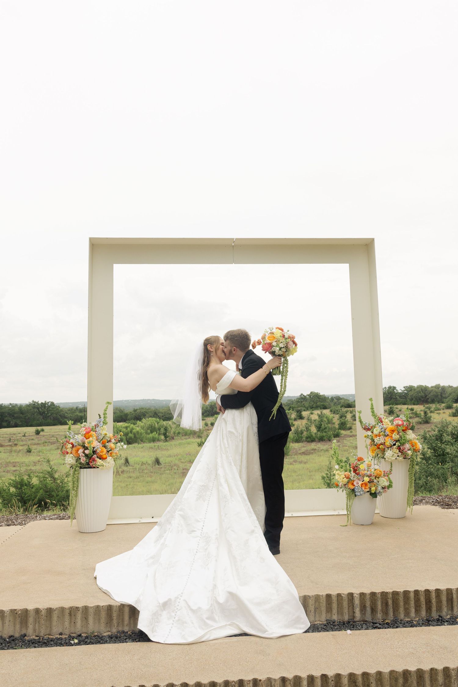 Bride and groom share first kiss under white square arch with orange floral arrangements at outdoor wedding ceremony.