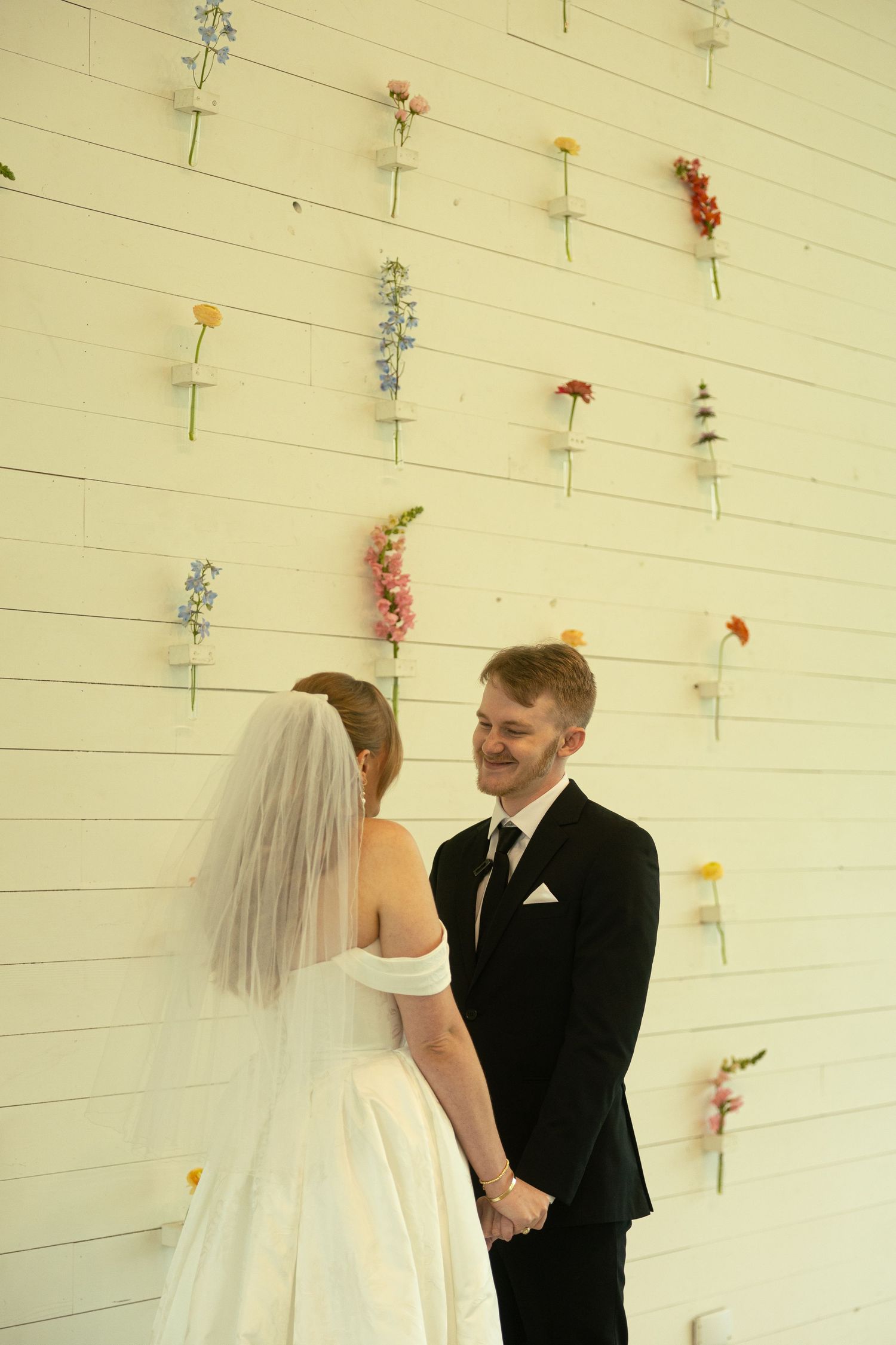 Bride and groom hold hands at wedding altar with individual flowers pinned to white shiplap wall backdrop.