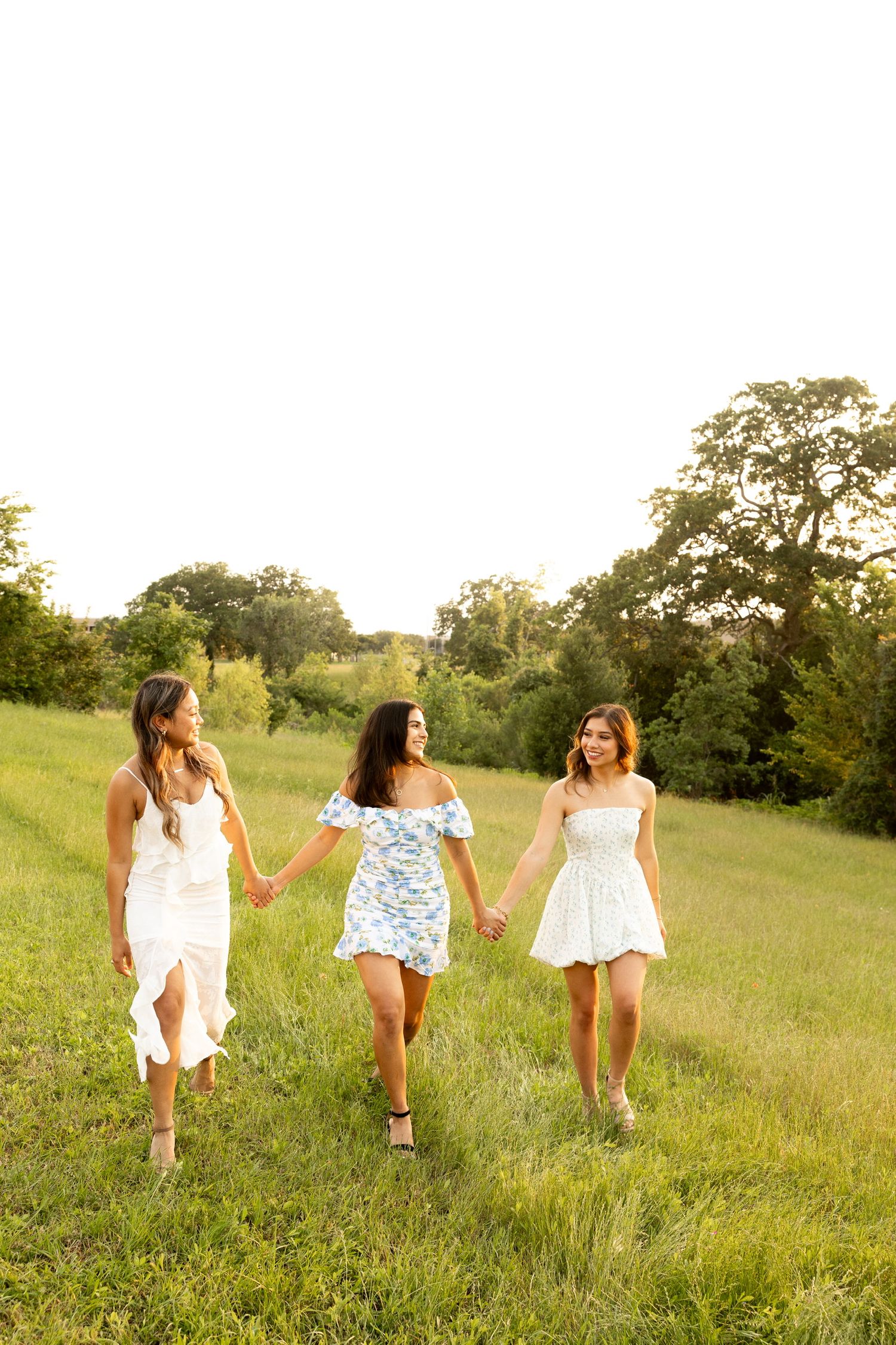Three friends hold hands while walking through a grassy hillside at sunset.