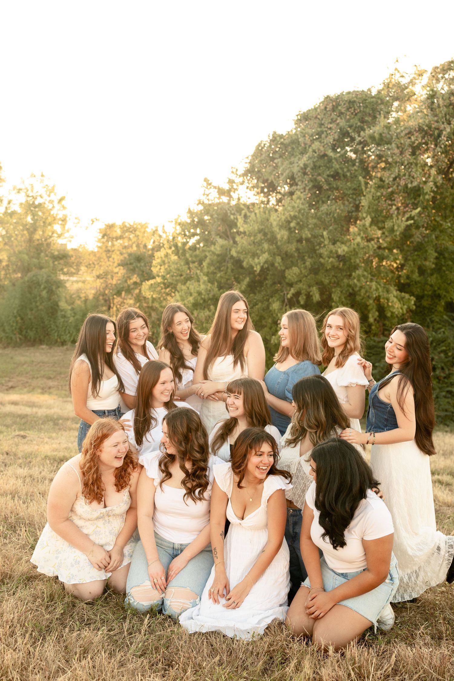 Large group of friends in white outfits sitting together on a grassy hill at sunset.