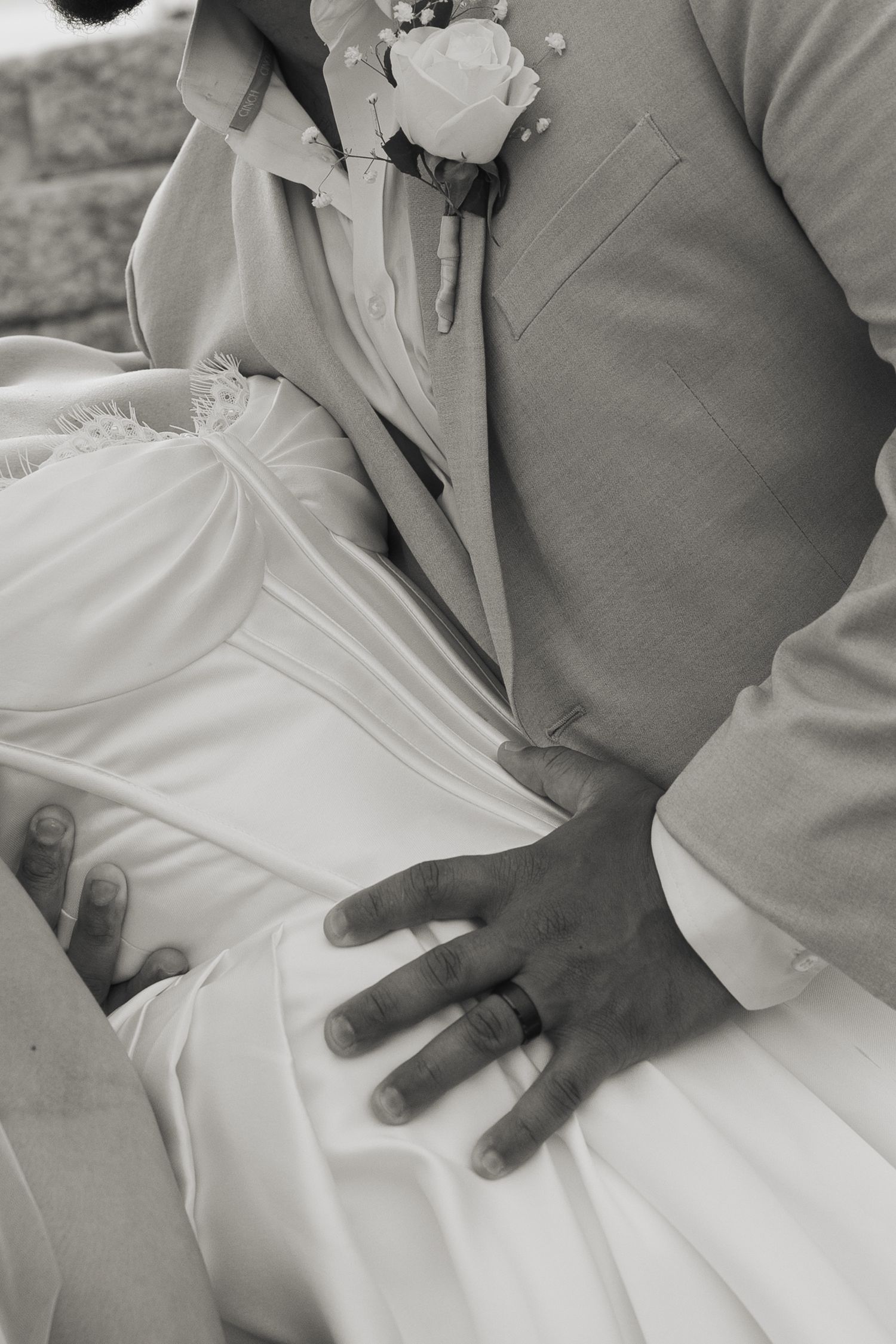Black and white close-up detail of a groom's hand on bride's white wedding dress with boutonniere visible.