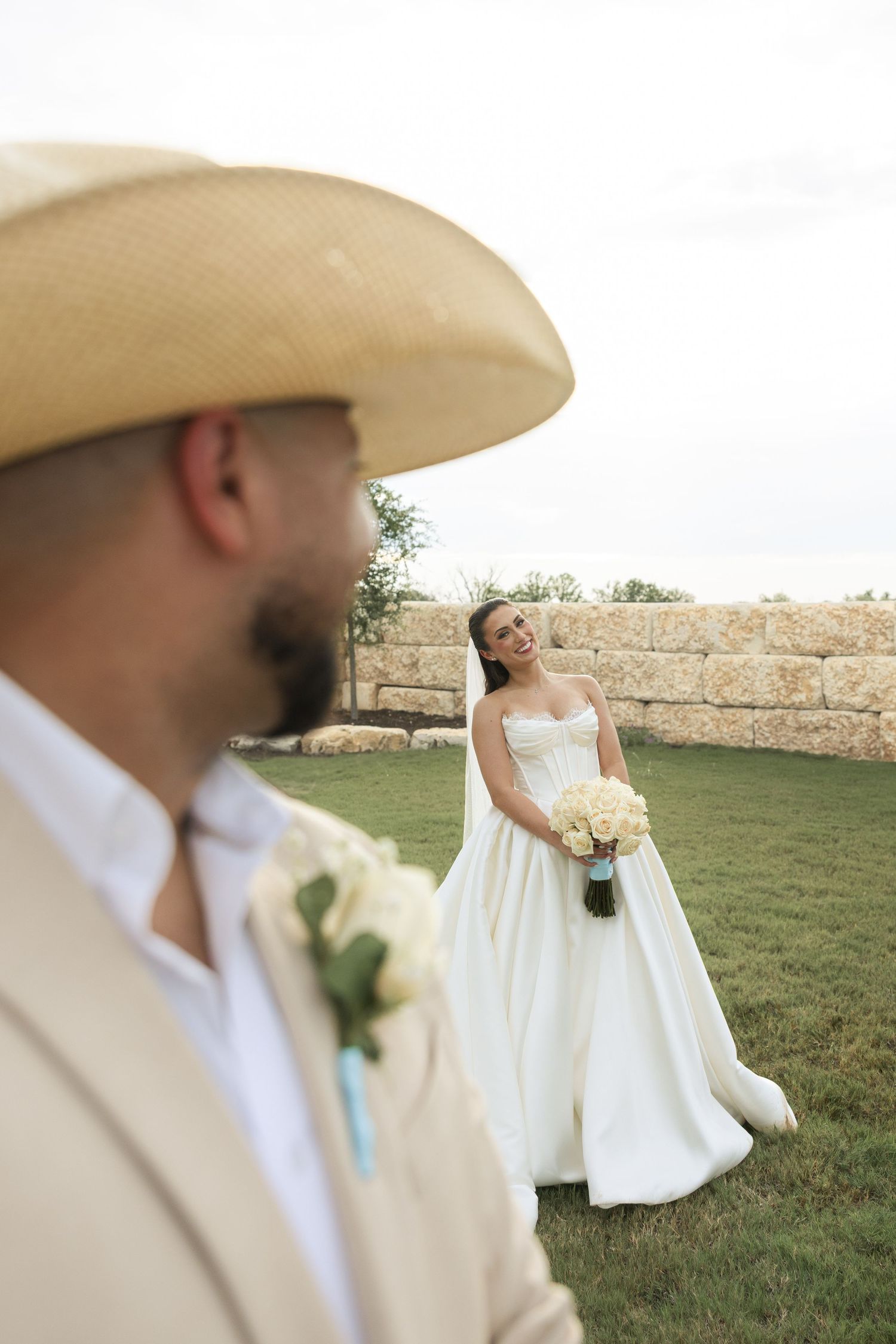 A bride in a white wedding gown holds a bouquet while standing on grass near a stone wall during an outdoor ceremony.