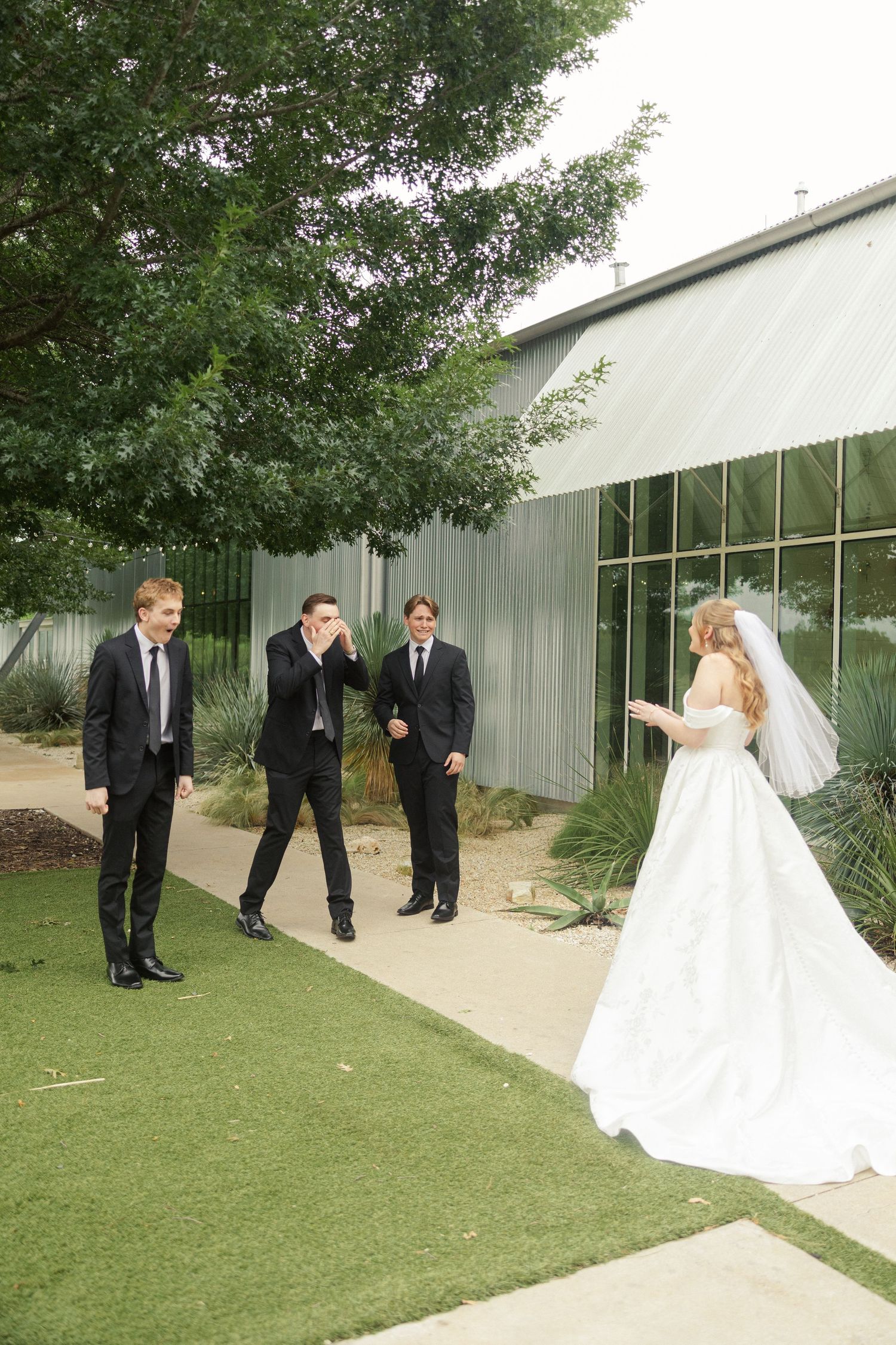 Bride in white gown and veil surprises emotional groomsmen in black suits outdoors at a modern wedding venue.