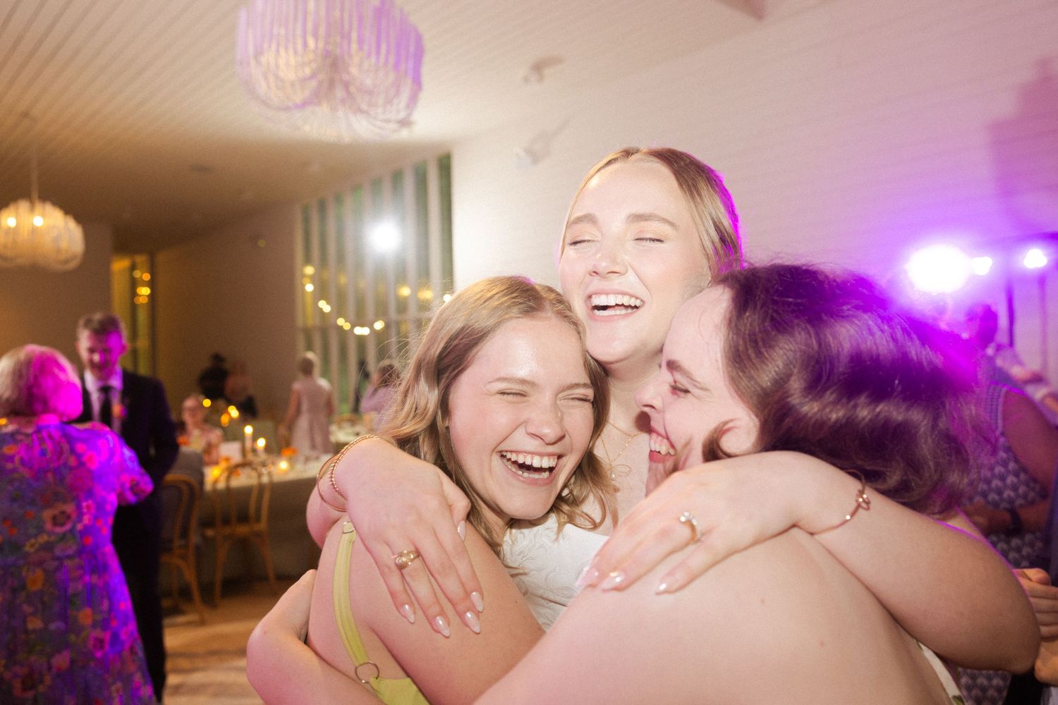 Three women laughing and hugging joyfully at a wedding reception with purple lighting.