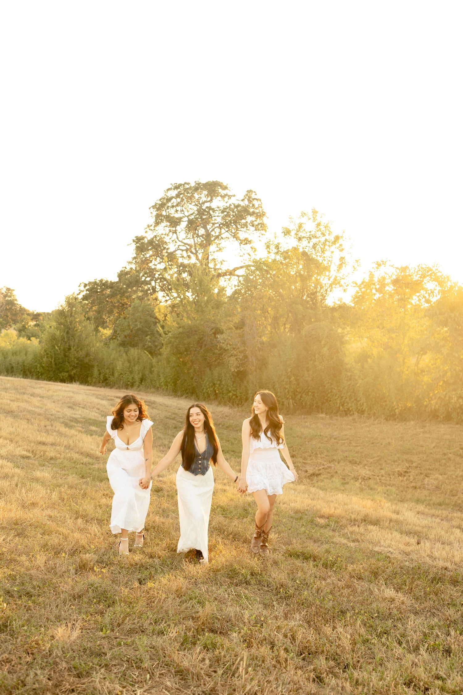 Three friends walking together in a sunlit meadow wearing white dresses during golden hour.
