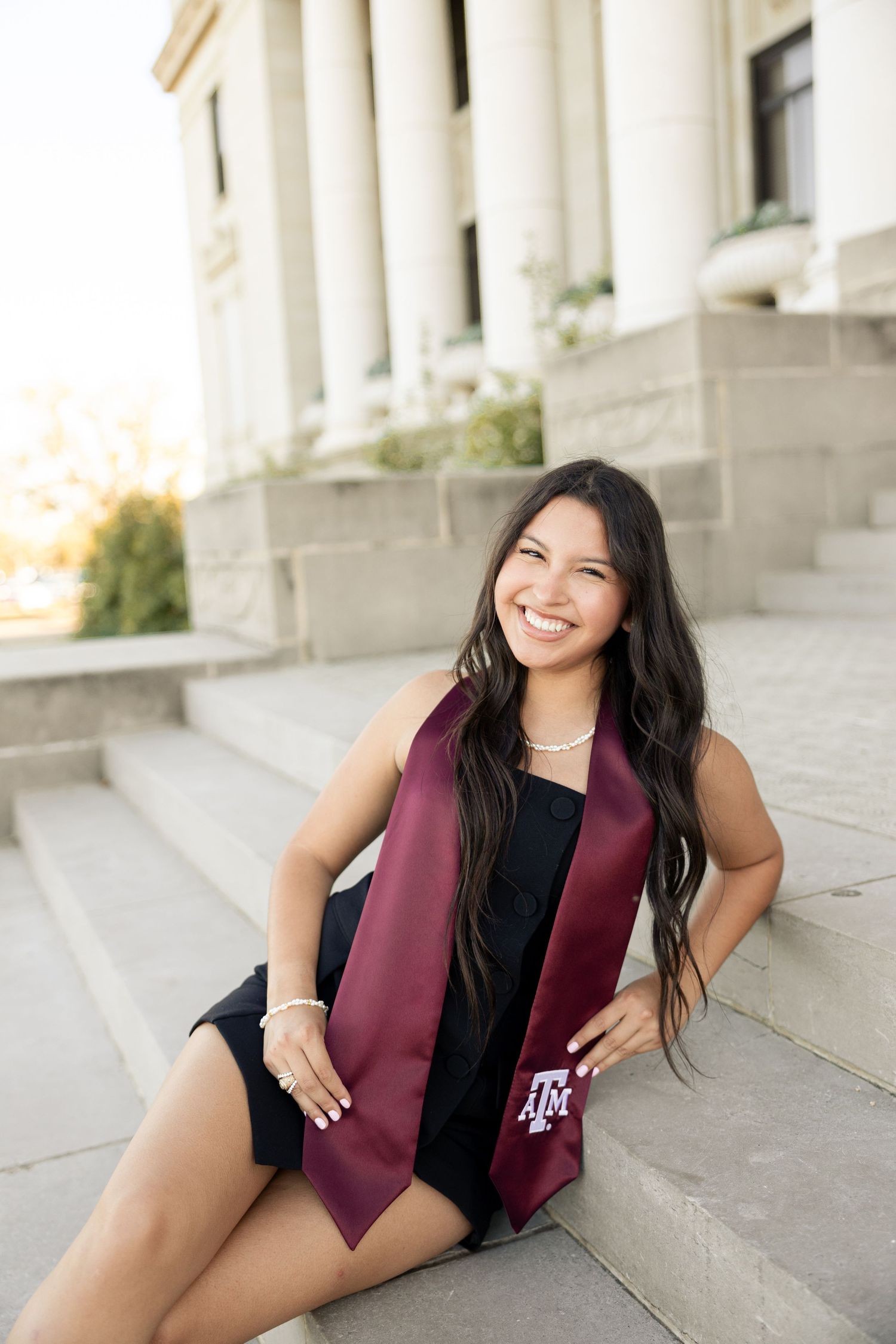 Smiling graduate in black dress and maroon stole poses on campus steps during sunset photo session.