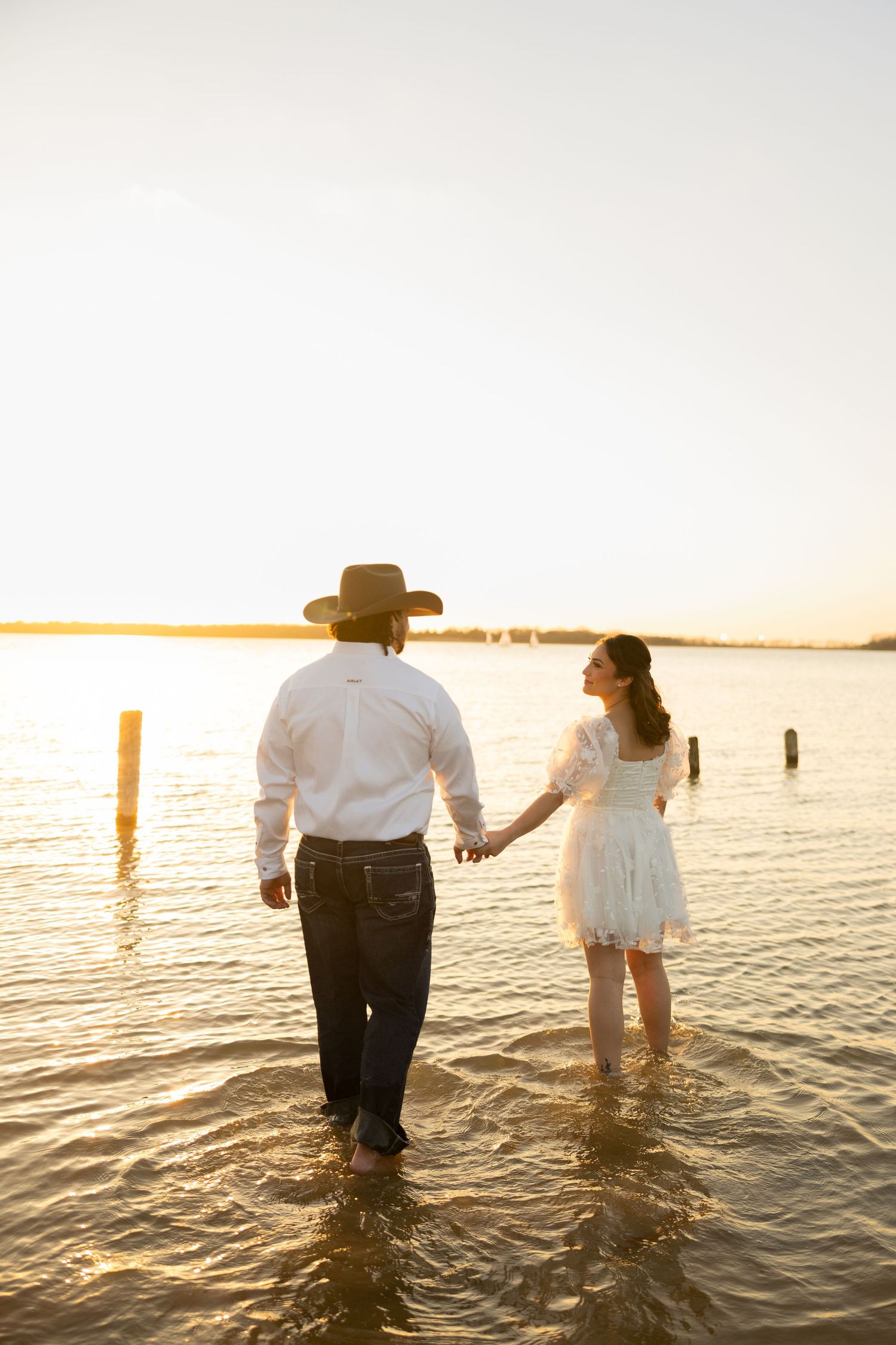 A couple holding hands wades in water during sunset near wooden posts.