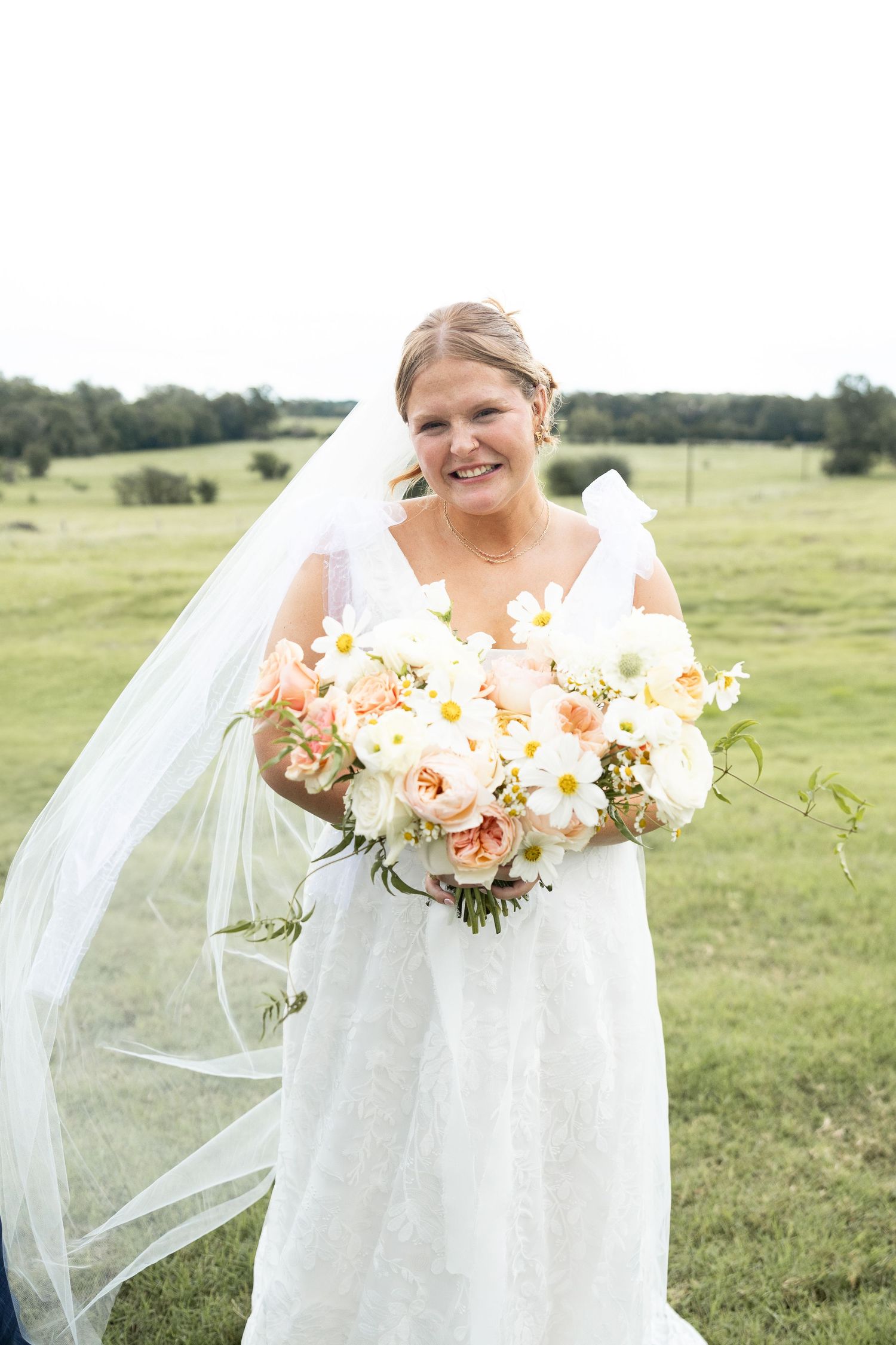A bride holds a beautiful bouquet of peach and white flowers against a green pastoral background.