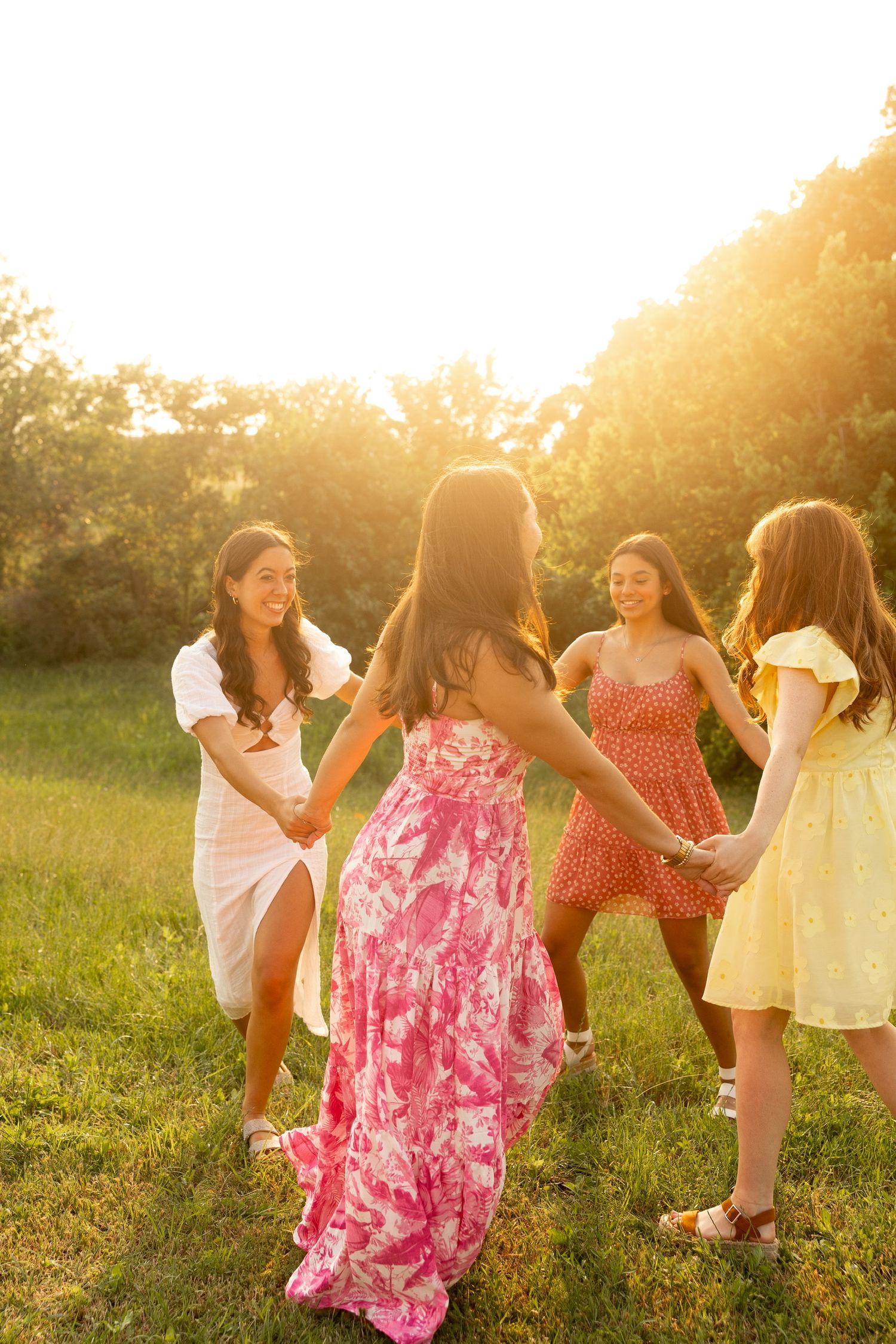 Friends in summer dresses dance together in a circle during golden hour in a grassy field.