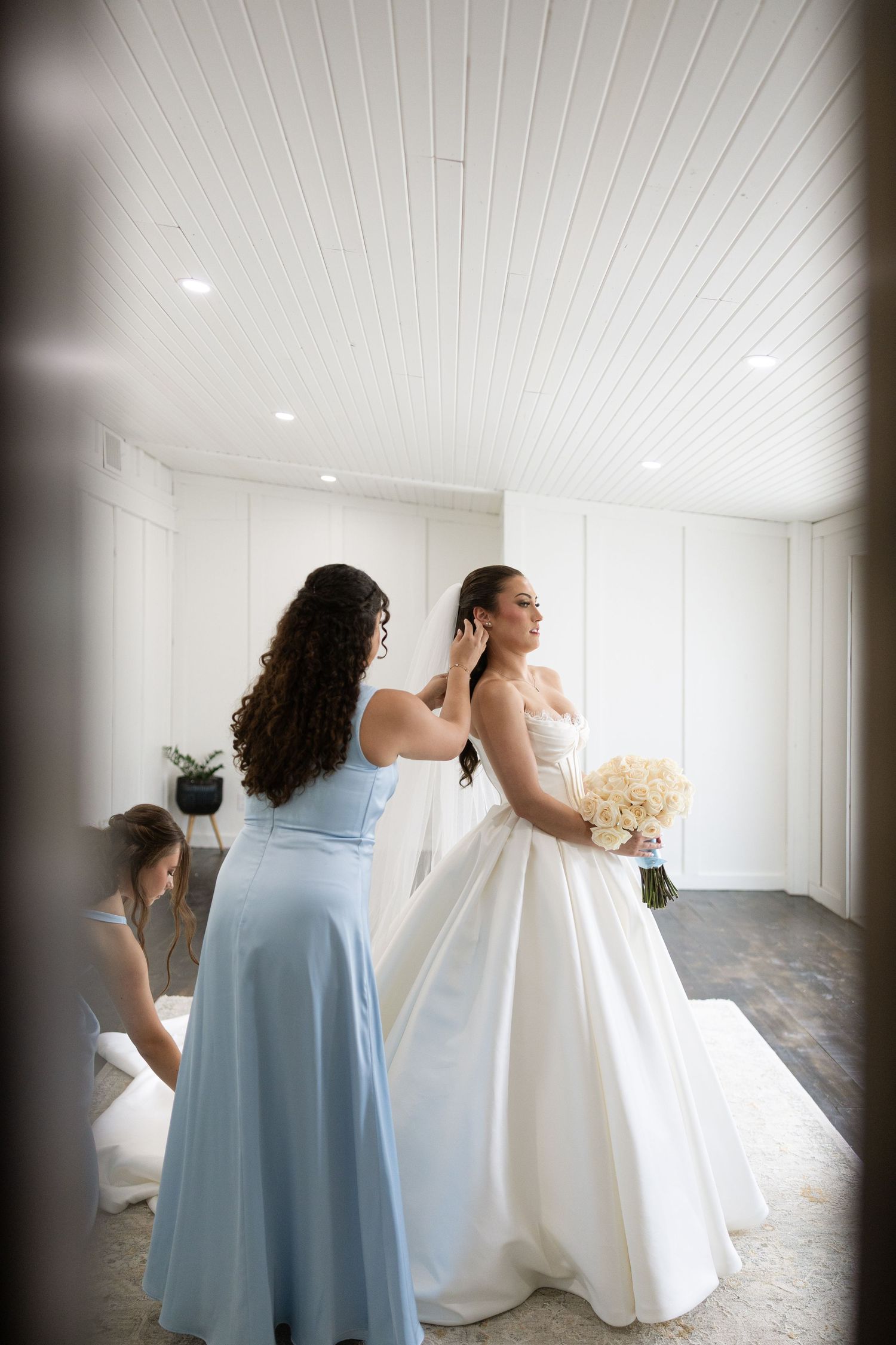 A bride in white gown poses with bridesmaid in light blue dress in an elegant white room.