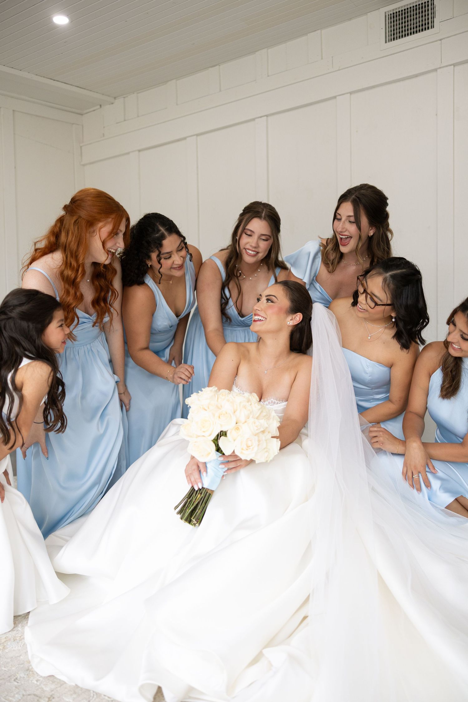 Bride in white wedding gown surrounded by bridesmaids wearing light blue dresses sharing a joyful moment together.