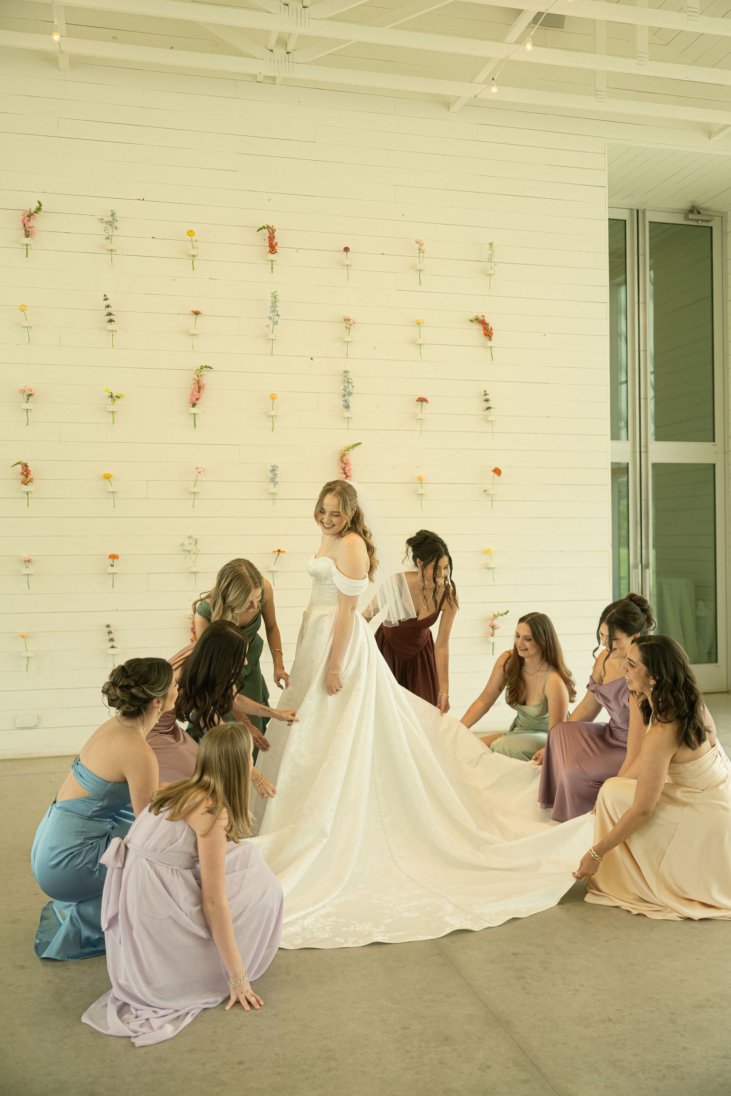 Bride in white gown surrounded by bridesmaids in colorful dresses arranging her train before wedding ceremony.