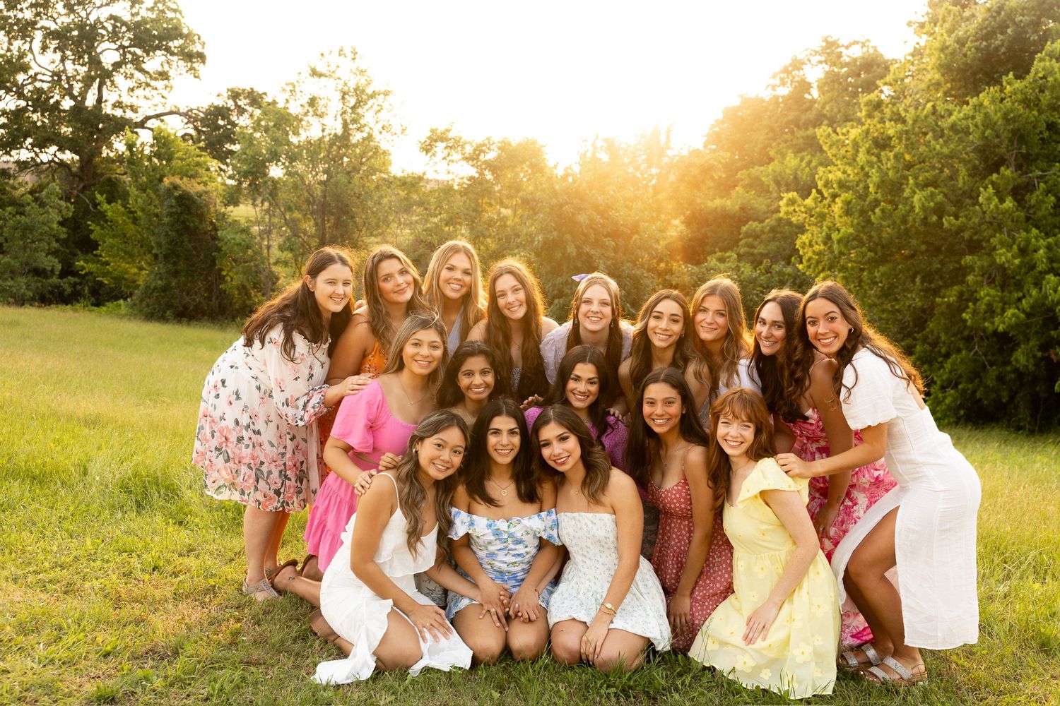Group wearing colorful summer dresses poses together in a grassy field at sunset.